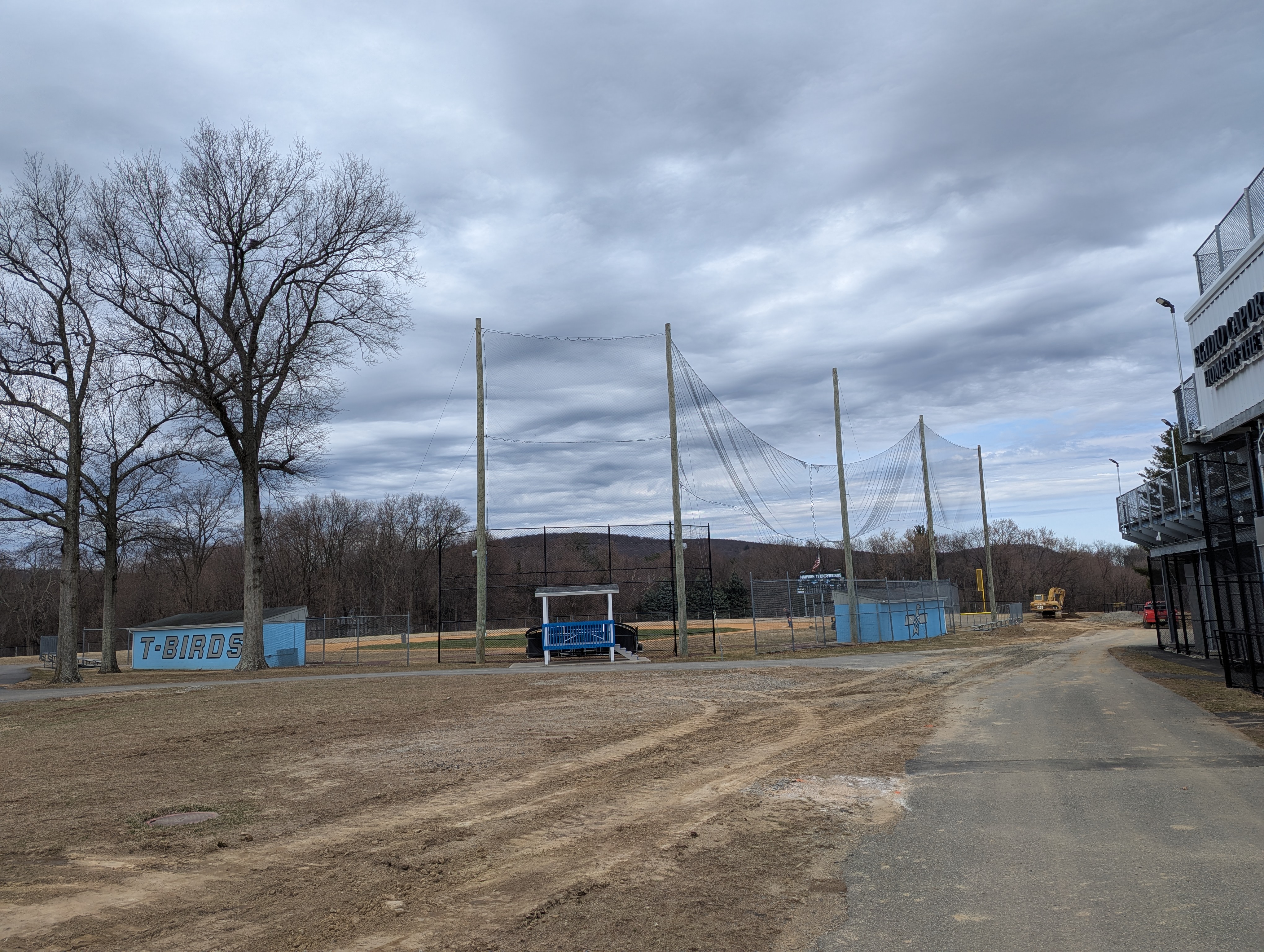 Bare trees and a dirt path are in the foreground, while a driving range with nets and a building are in the background under a cloudy sky.