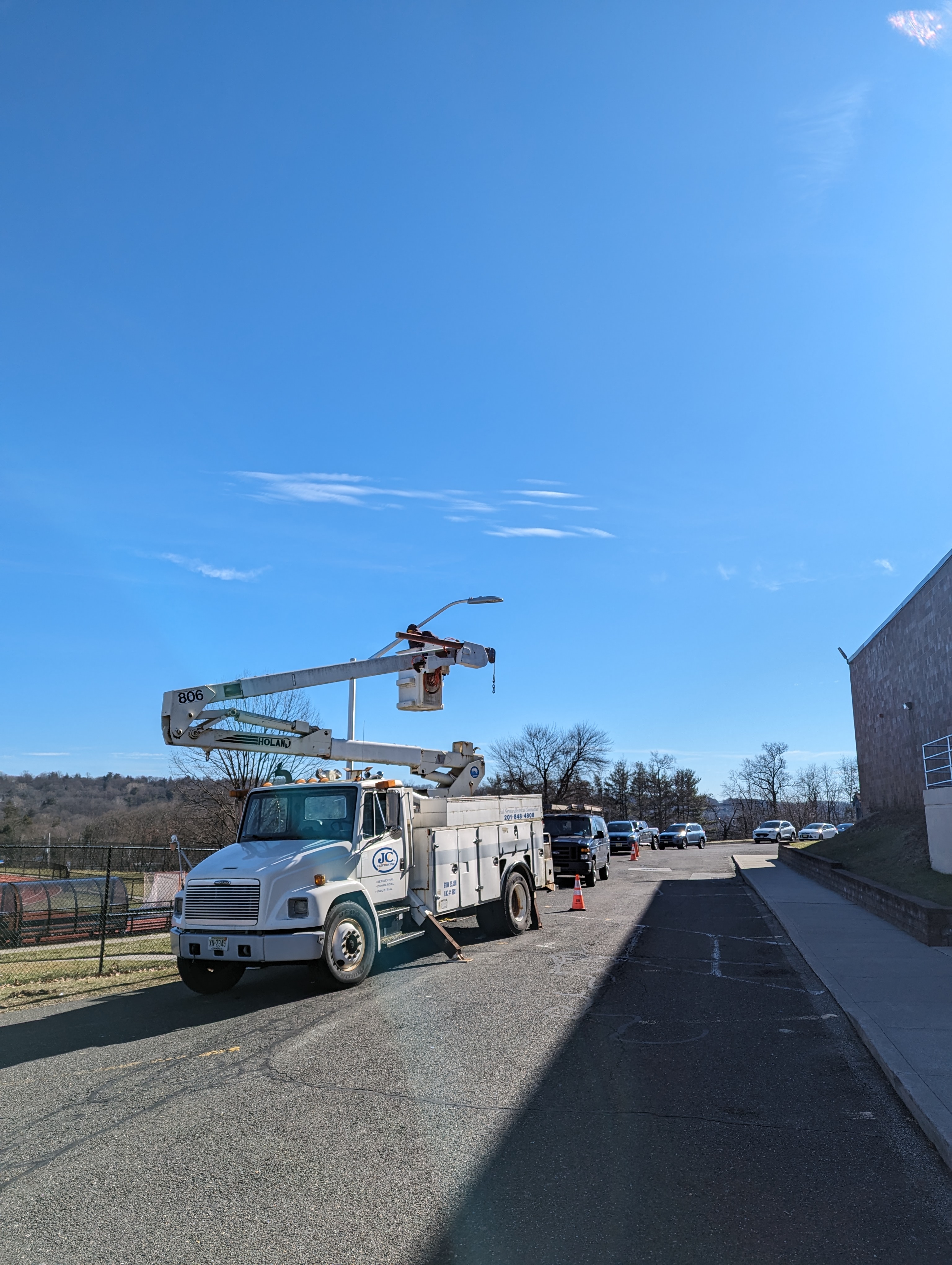 A bucket truck is parked on a paved road in the foreground, with a line of vehicles and a building visible in the background under a clear blue sky.