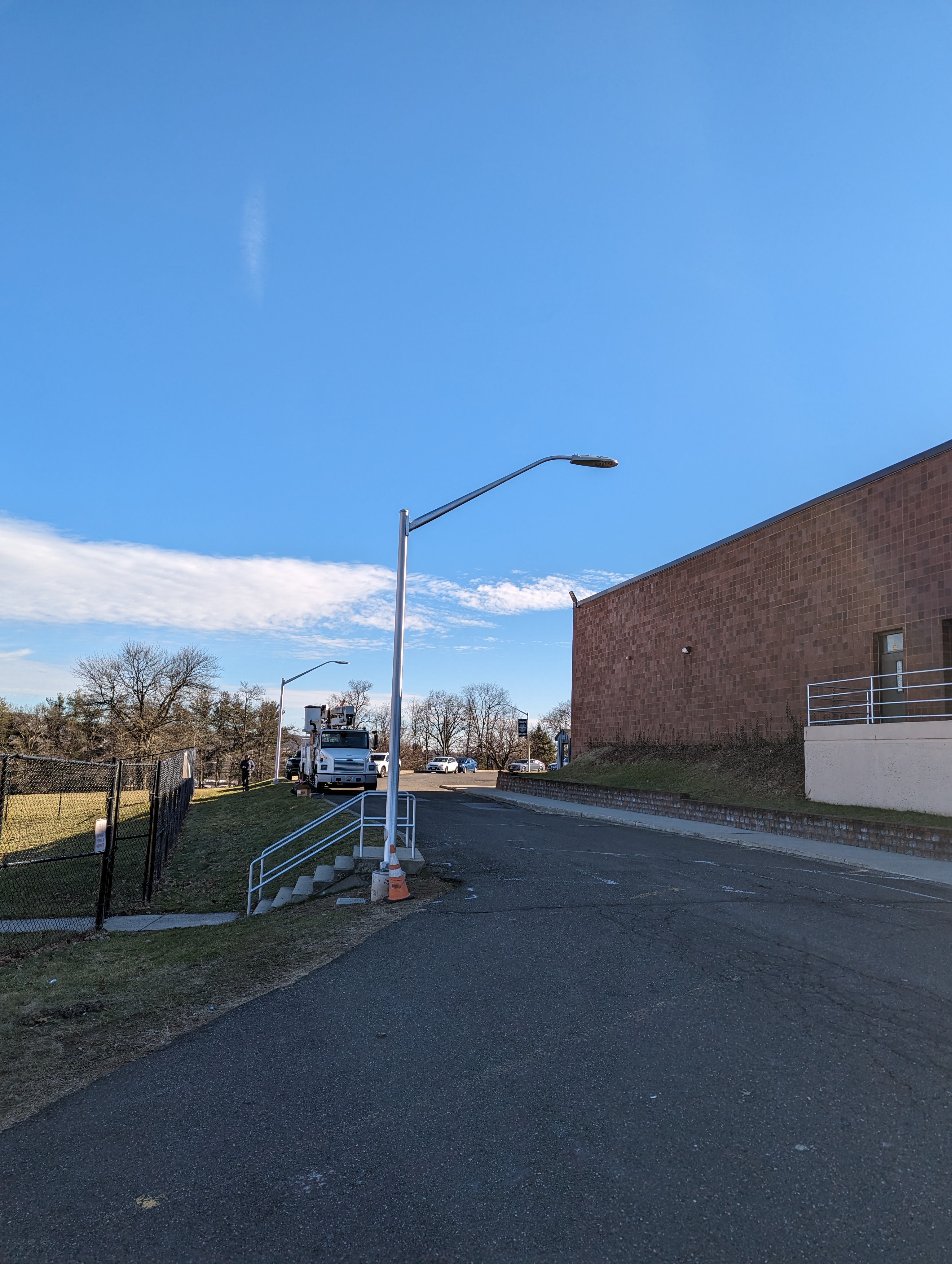 A tall street lamp stands in the foreground on a paved path leading towards a brick building, with a utility truck and trees visible in the background under a clear blue sky.