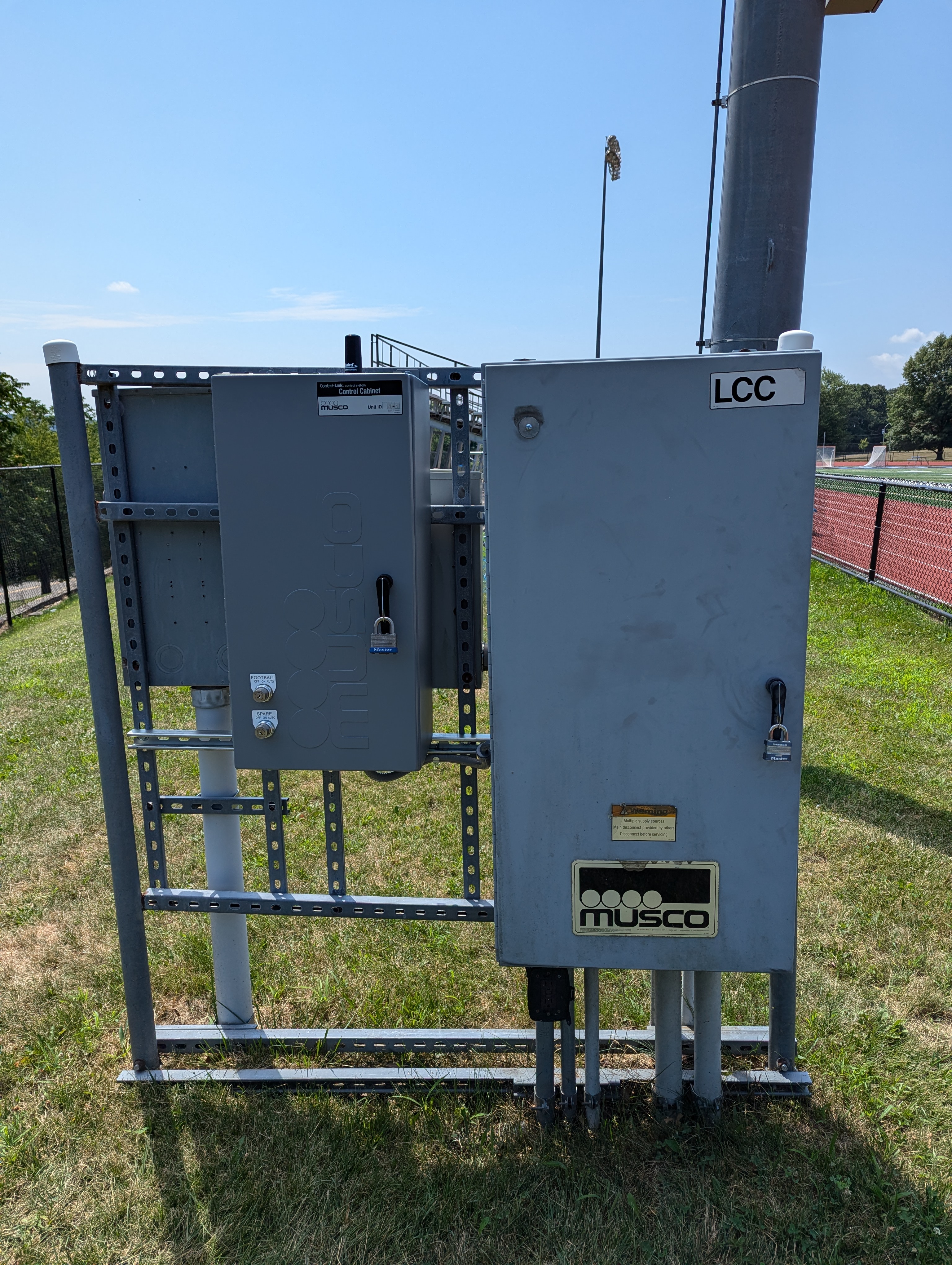 Two gray electrical boxes stand on a metal frame in the foreground, with a track and trees visible in the background under a clear blue sky.