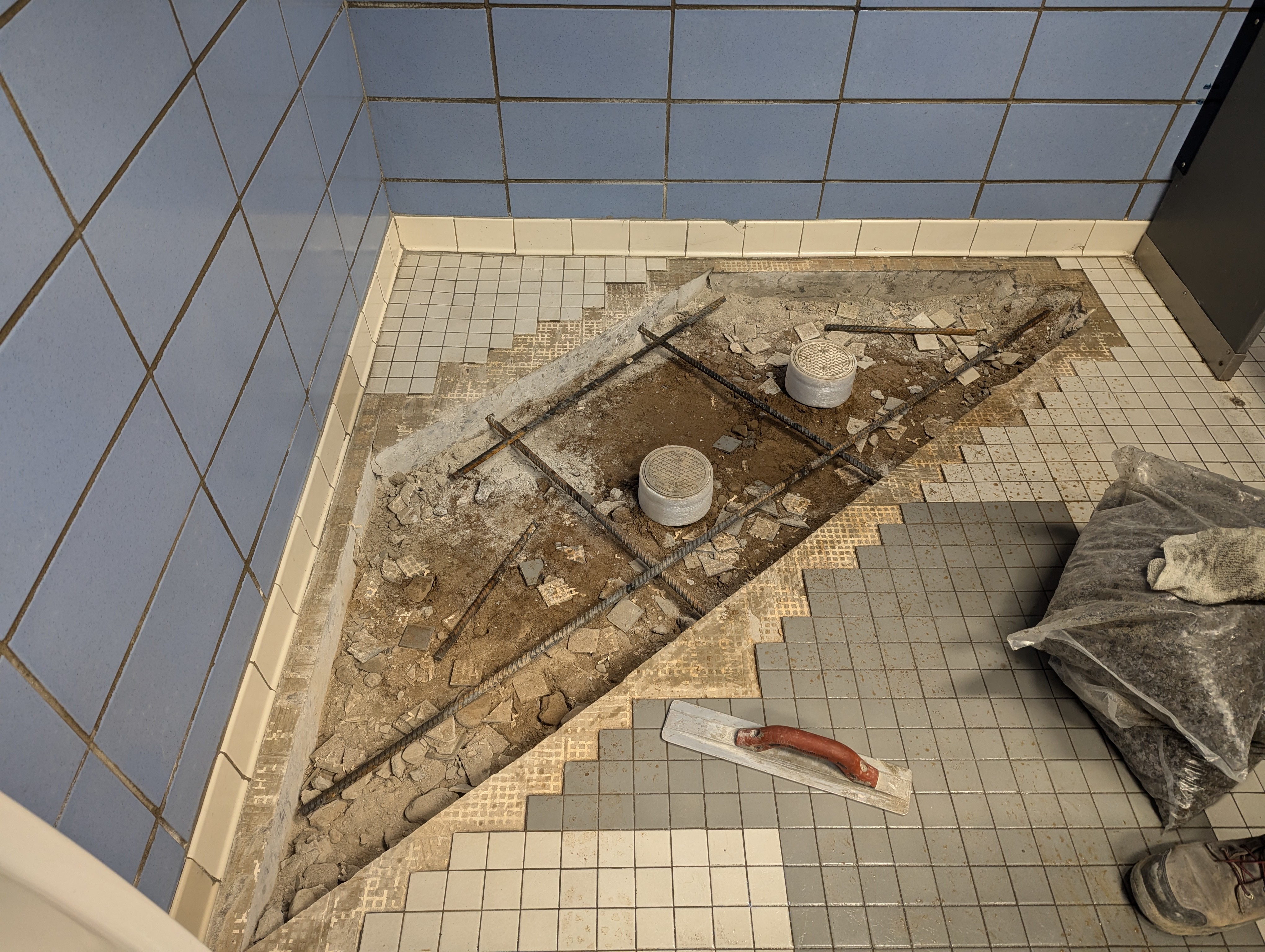 In the foreground, exposed rebar and concrete are visible in a damaged tiled floor, while the background features light blue tiled walls and a dark partition.