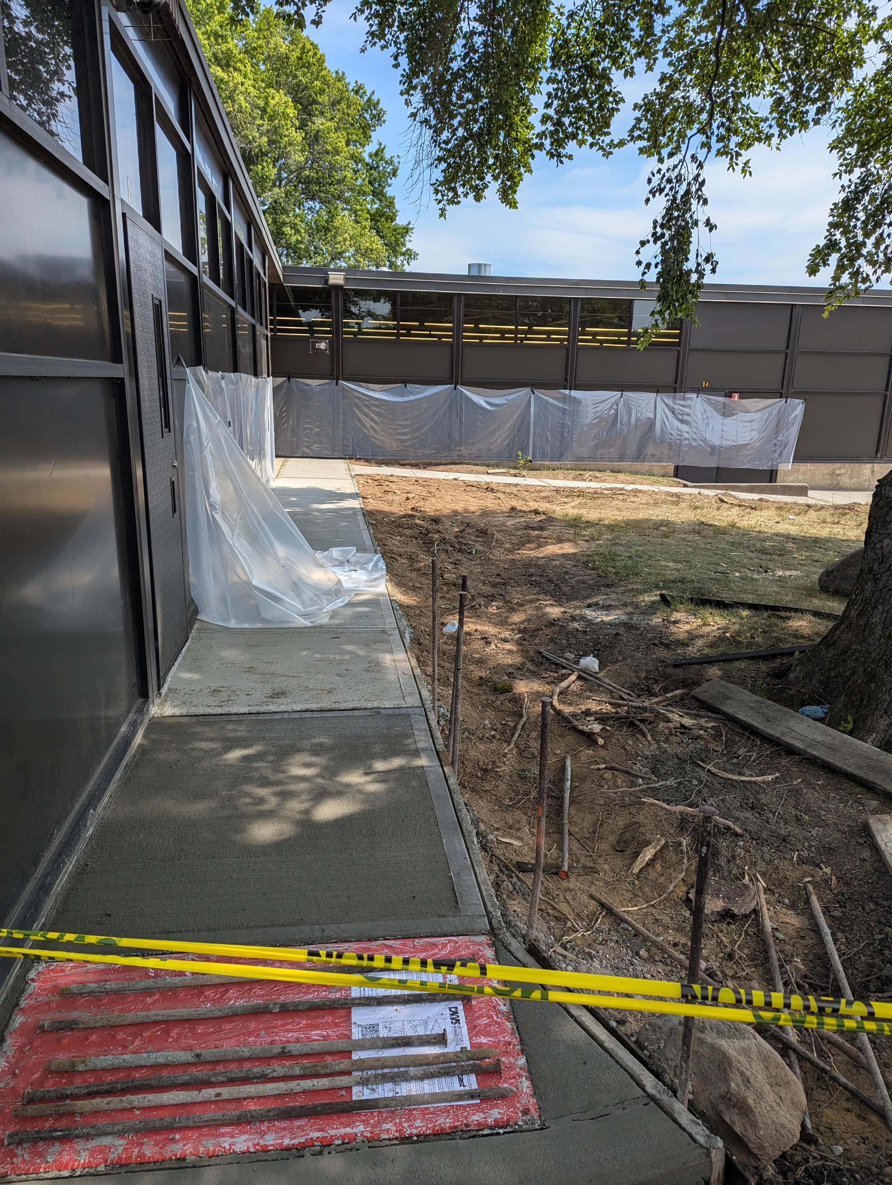 Freshly poured concrete forms a new walkway in the foreground, leading past a building with large windows, while the background shows a construction site with protective sheeting and trees.