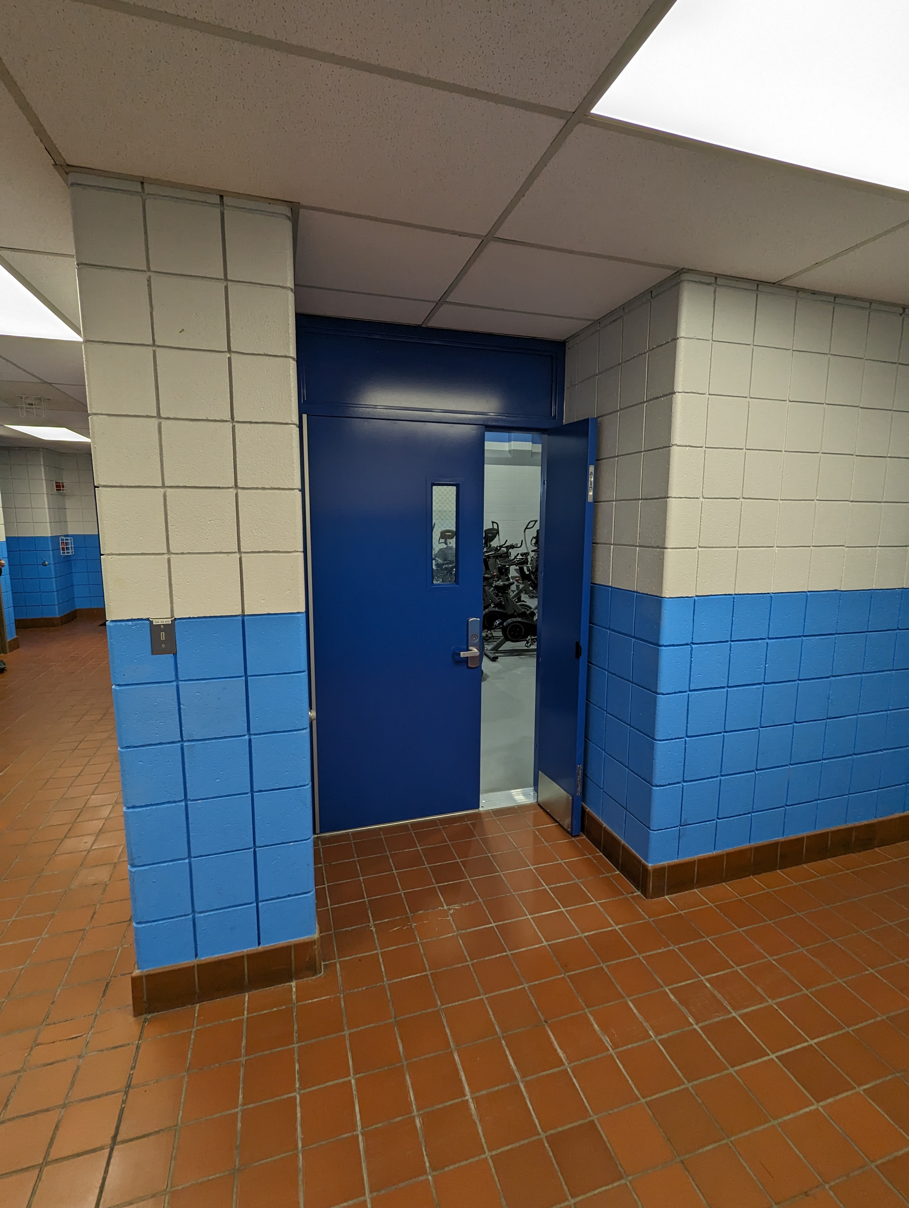 A blue door stands ajar in a tiled hallway with a tiled floor in the foreground and a glimpse of gym equipment in the background.