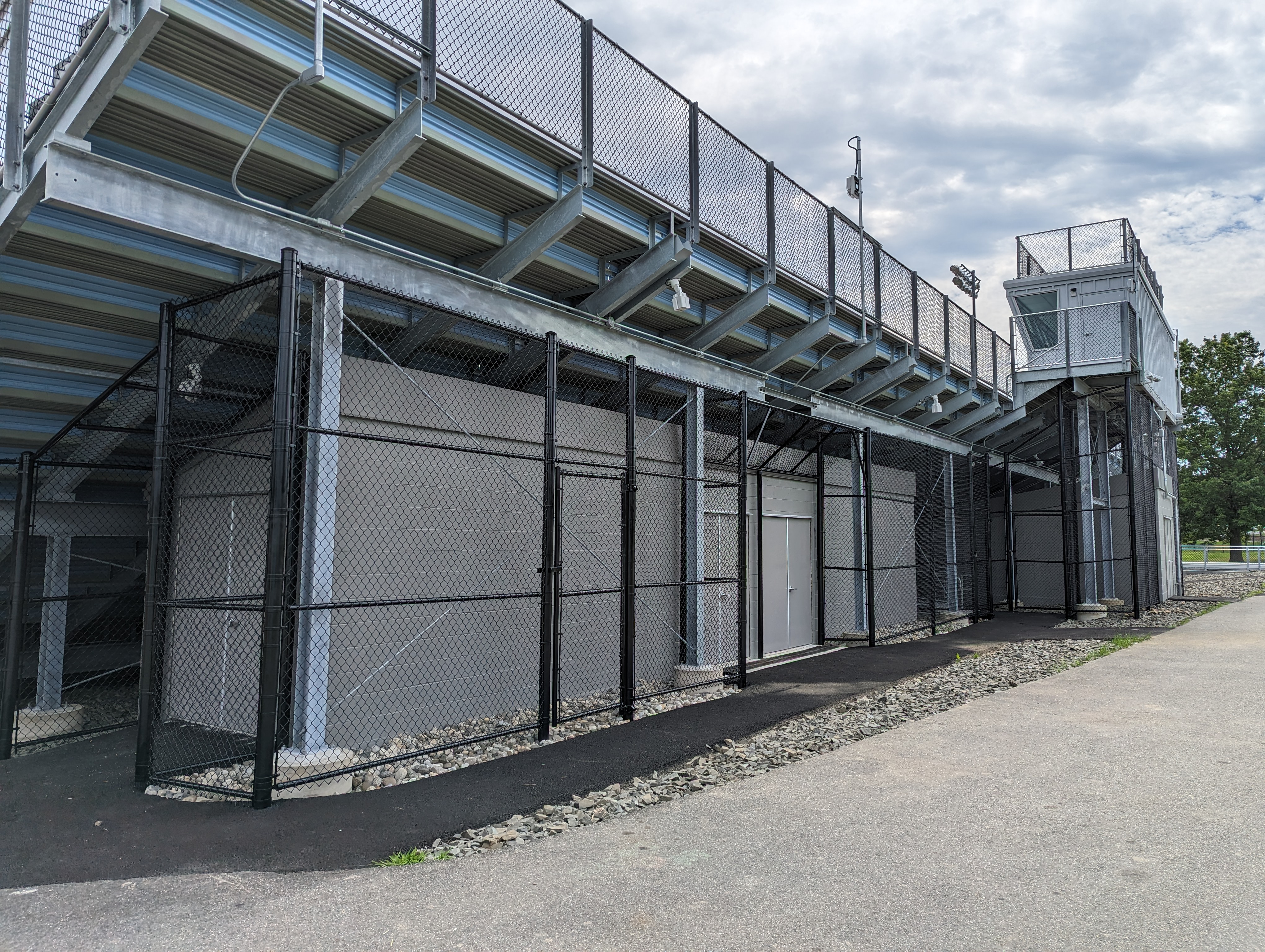 A paved walkway leads past a chain-link fence and storage area to the bleachers of an empty stadium under a cloudy sky.