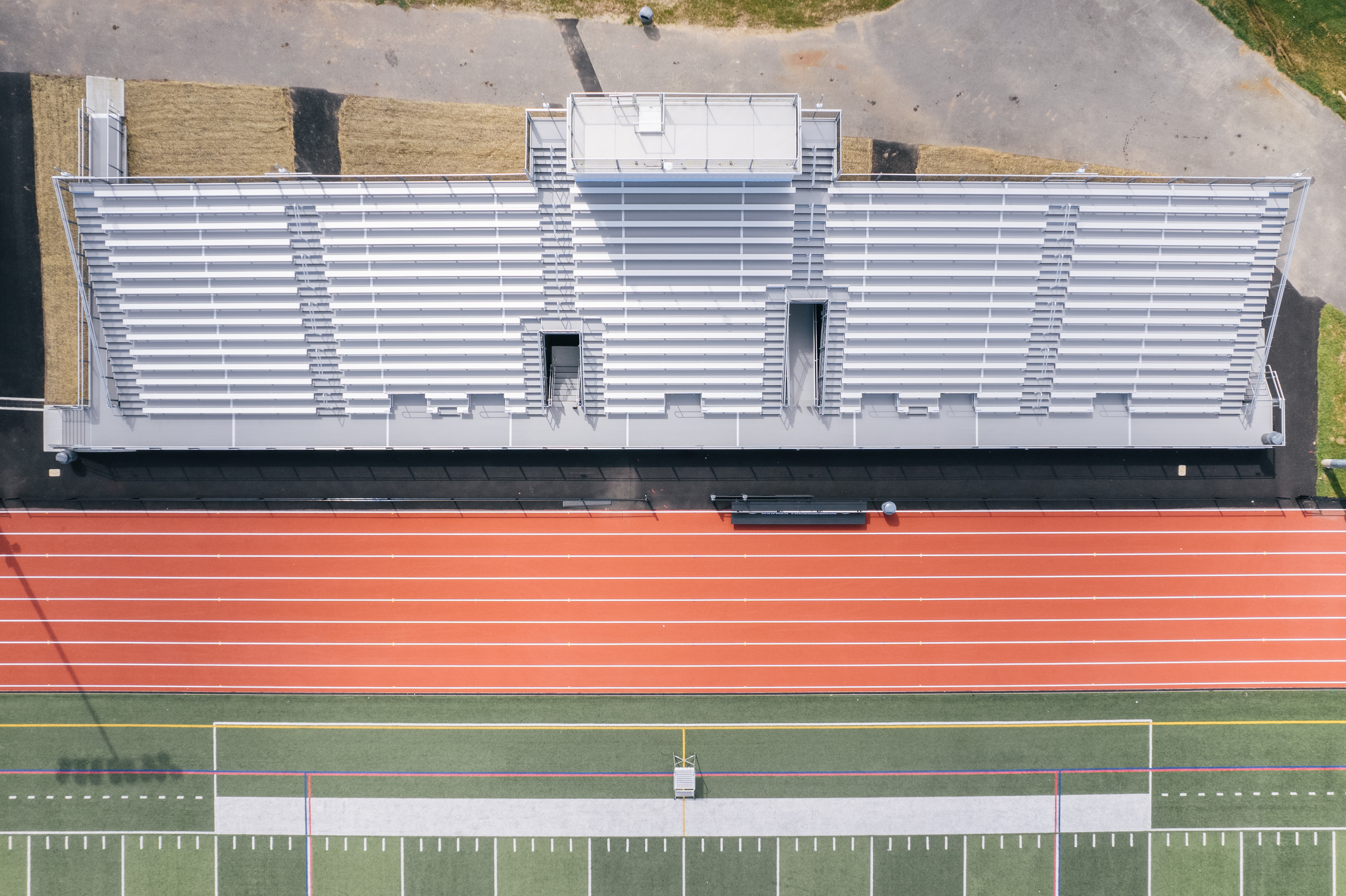 An aerial view captures a stadium's empty bleachers and track in the foreground, with a grassy field and a parking lot in the background.
