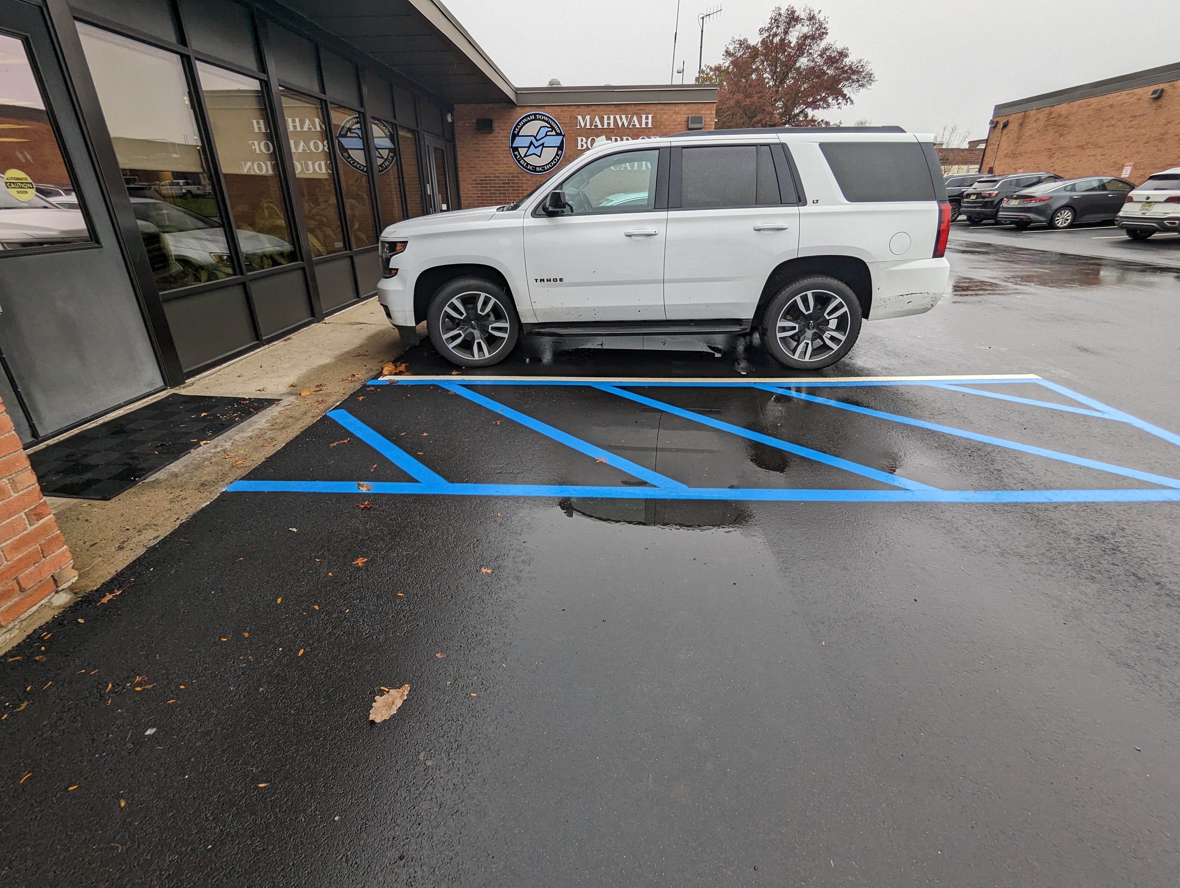 A white SUV is parked in a wet, blue-lined parking space in front of a brick building with "MAHWAH BOOKKEEPING" signage, while other cars are visible in the background.