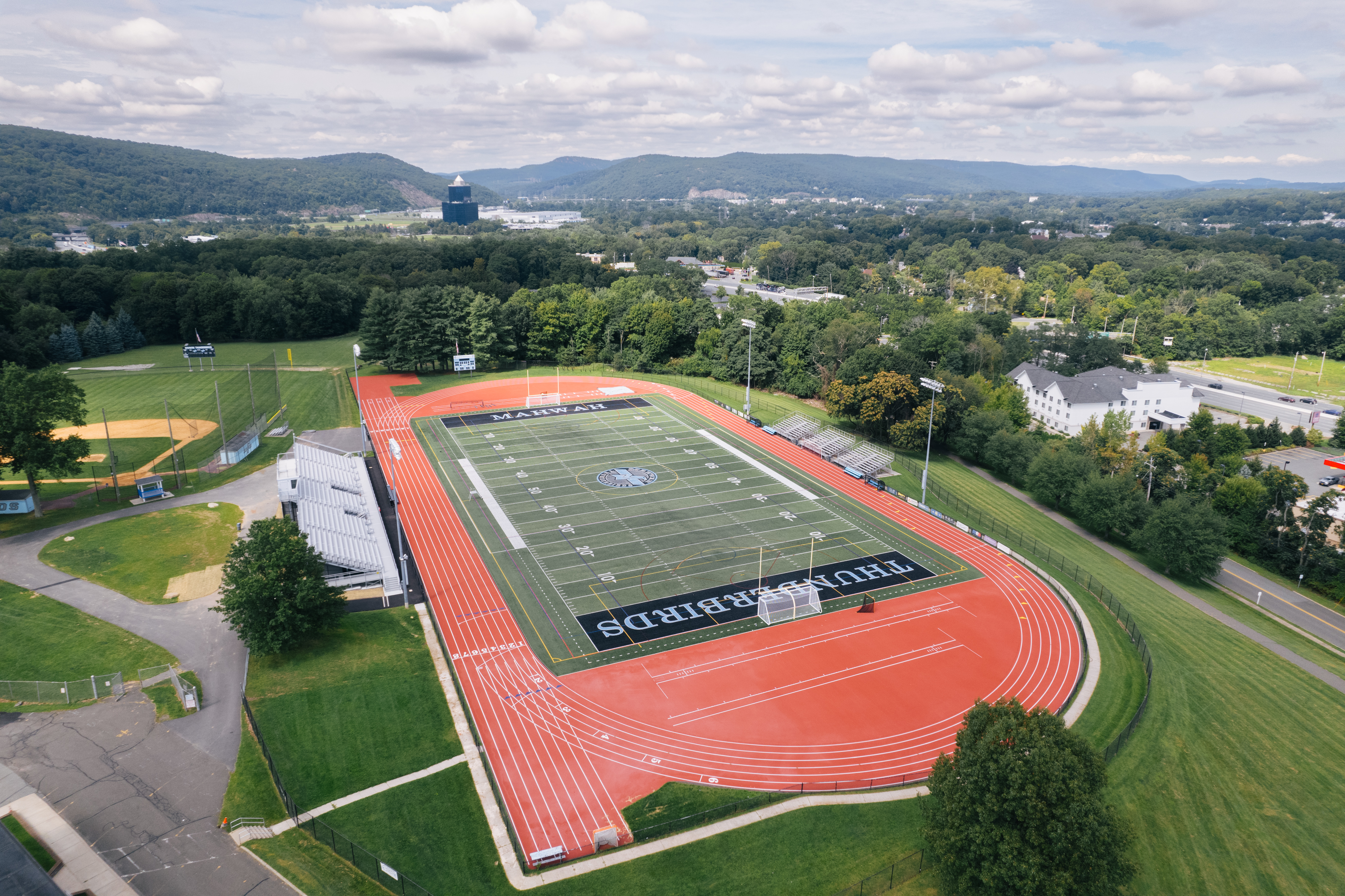 A vibrant red running track encircles a football field with "THUNDERBIRDS" written across it, set against a backdrop of lush green trees and distant hills.