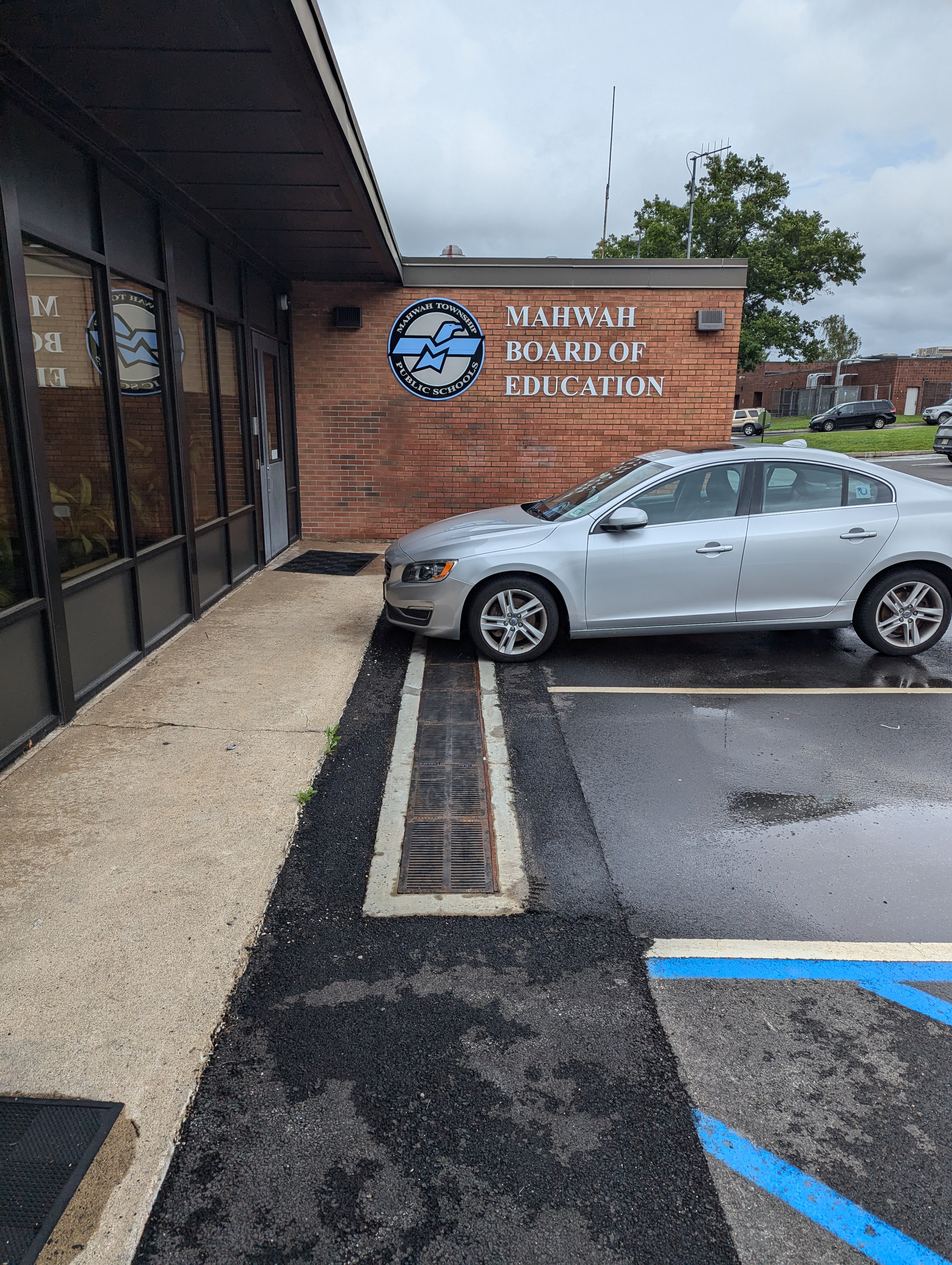 A silver car is parked in front of a brick building with "MAHWAH BOARD OF EDUCATION" signage, with a sidewalk and some greenery in the foreground.