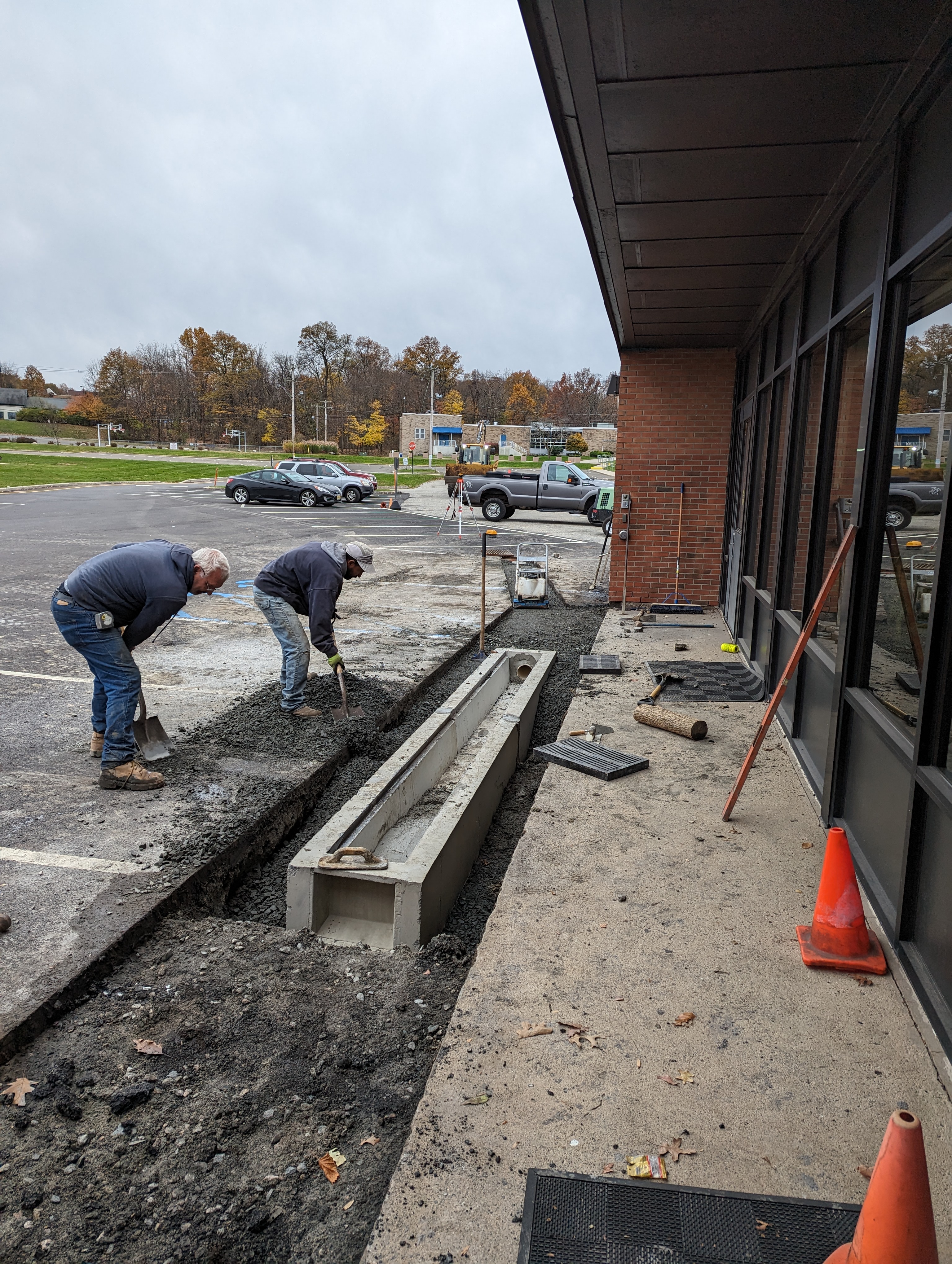 Two workers dig a trench in a parking lot next to a building, with cars and trees visible in the background.