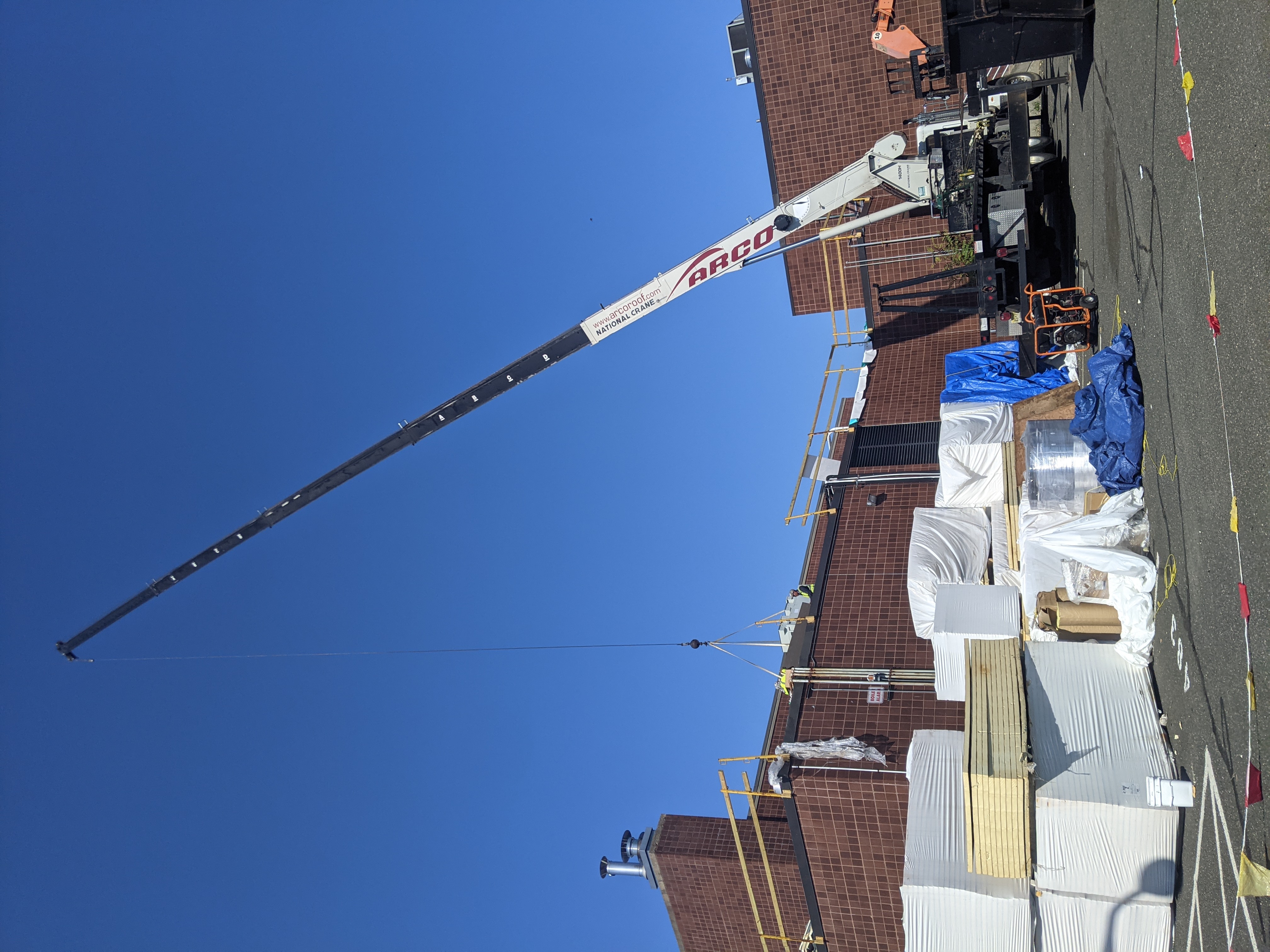 A crane lifts materials over stacks of insulation and building supplies in front of a brick building under a clear blue sky.