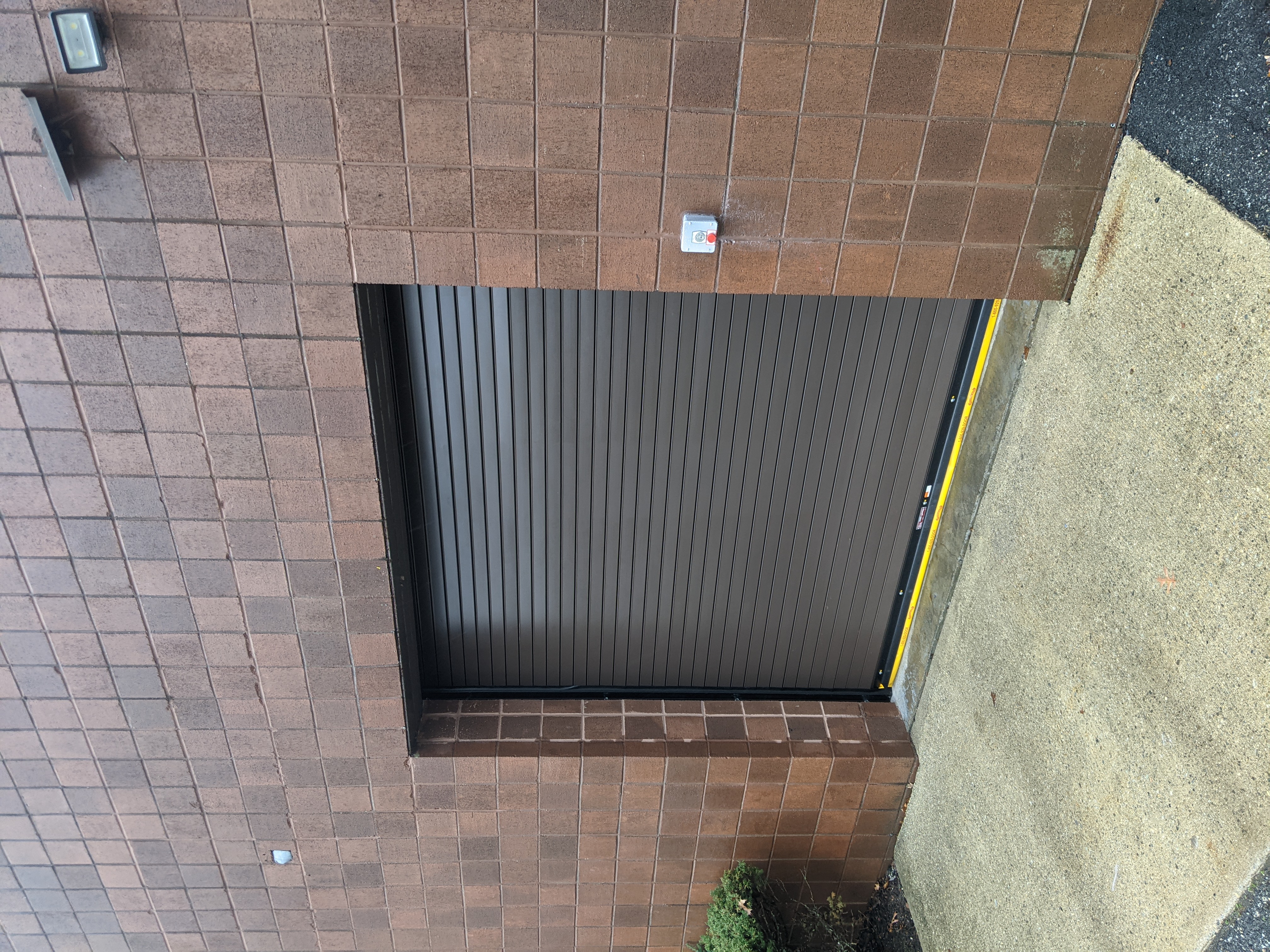 A closed, dark brown roll-up door is set into a brown brick wall, with a concrete walkway in the foreground.