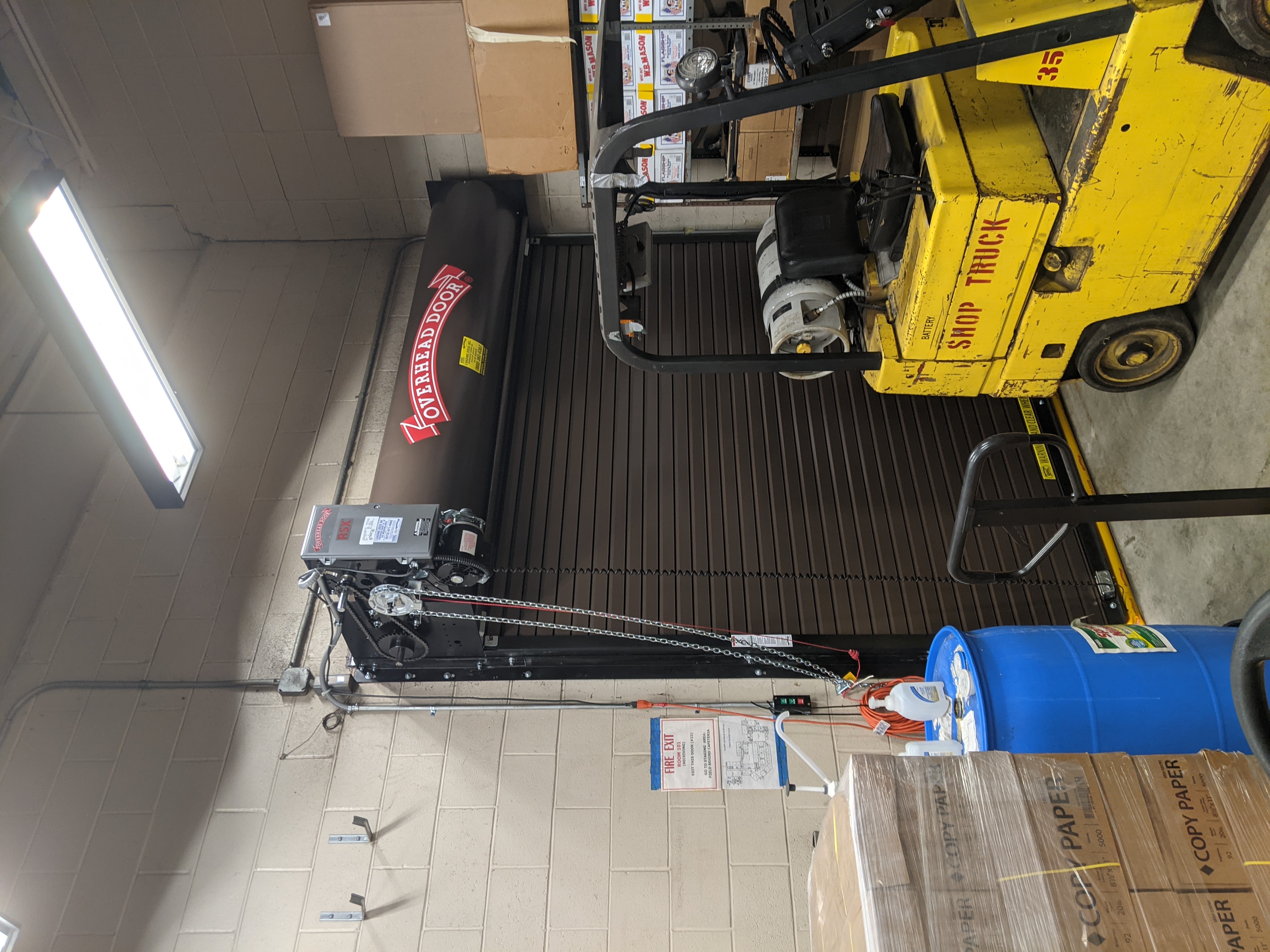 A yellow forklift is parked in front of a closed brown roll-up door, with stacks of paper and a blue barrel in the foreground and a light fixture on the ceiling in the background.