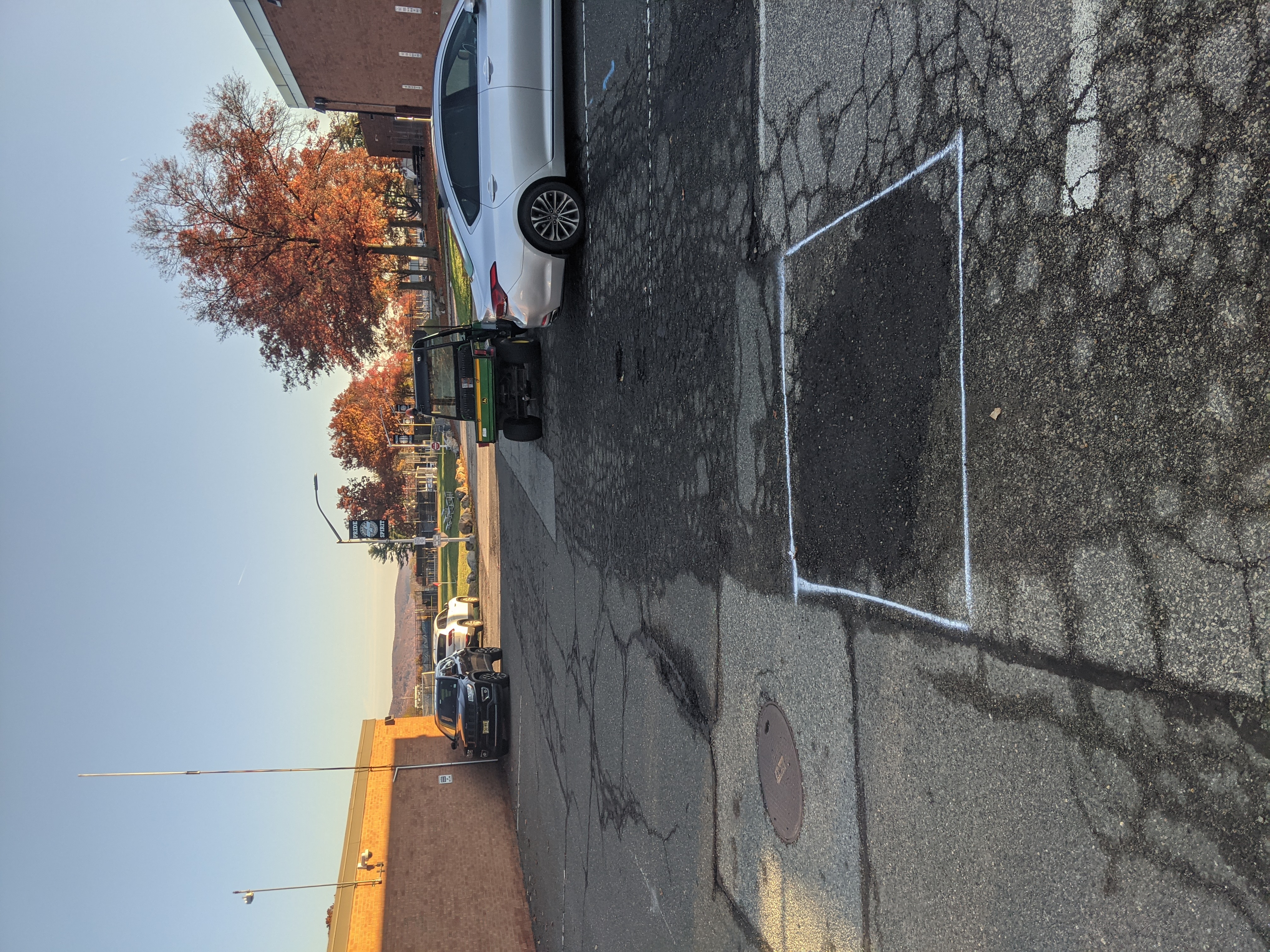 A patch of cracked asphalt marked with white lines is in the foreground, while a utility vehicle and cars are parked in the background under a clear sky with autumn trees.