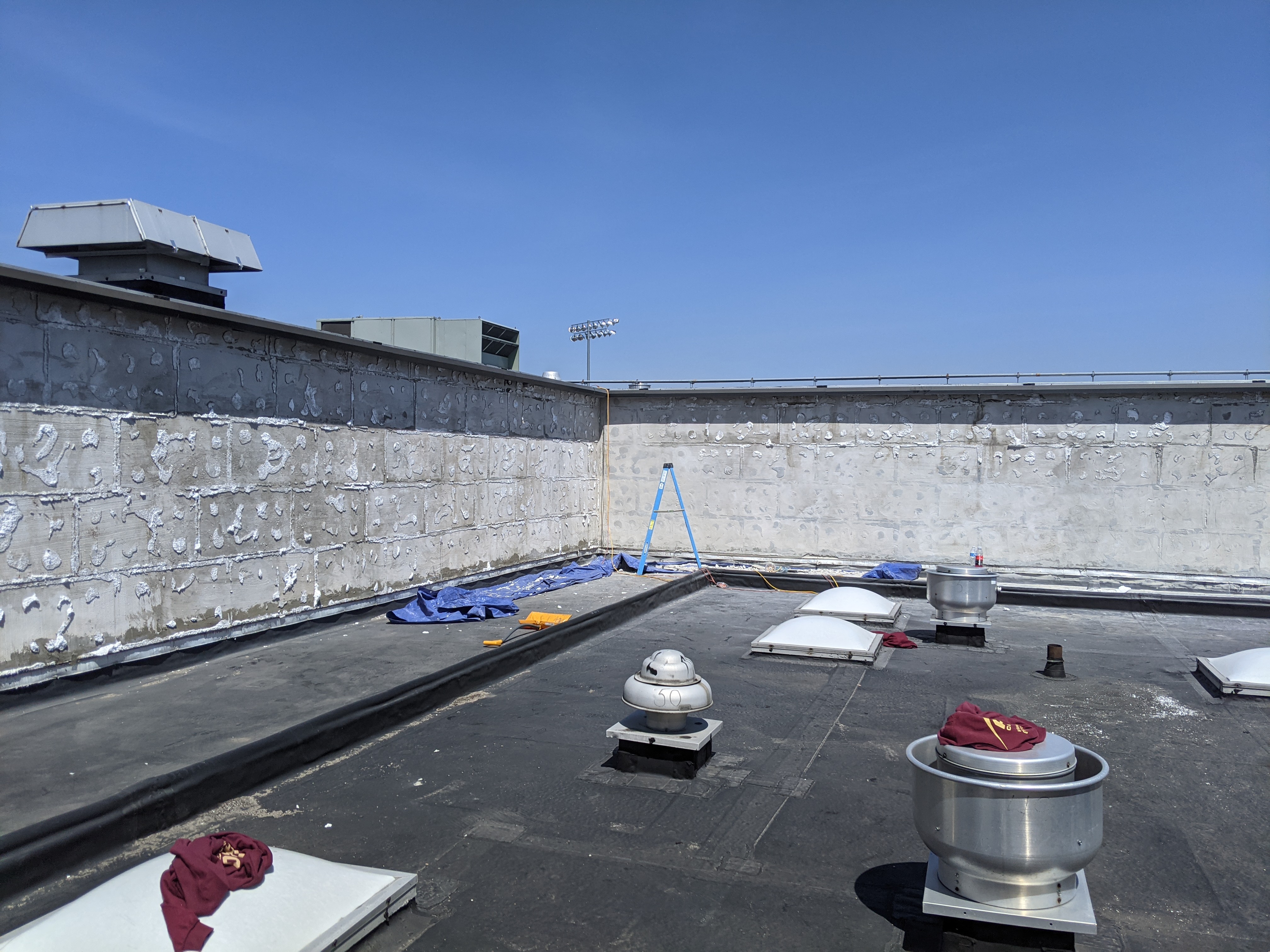 A flat, dark rooftop with several vents and skylights in the foreground is bordered by a weathered concrete wall and a clear blue sky in the background.