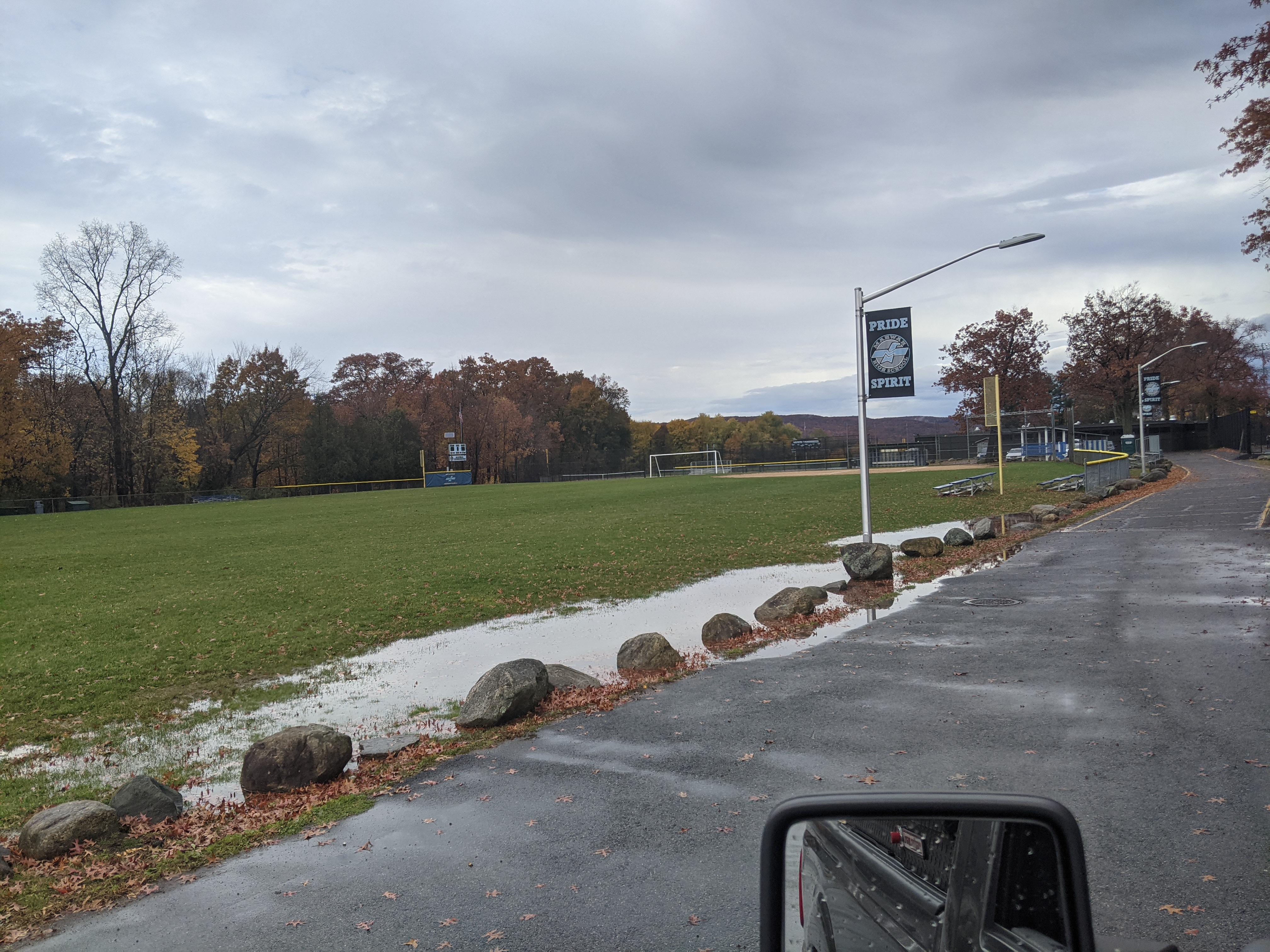 A wet asphalt road with rocks and fallen leaves in the foreground leads to a grassy field with trees and sports facilities in the background under a cloudy sky.