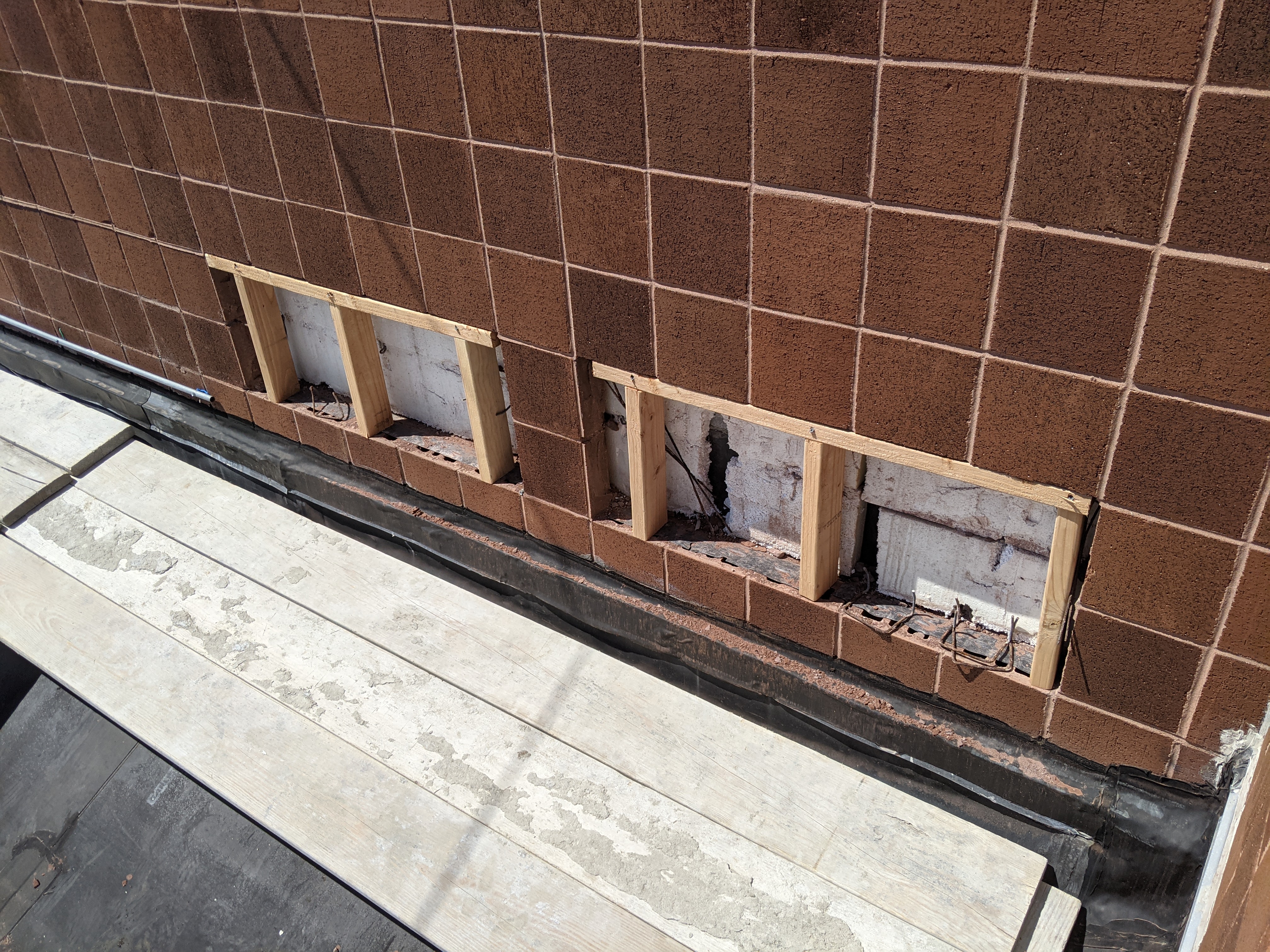 Brown tiled wall with three open, unfinished window frames in the background, and a section of light-colored planks and dark roofing material in the foreground.