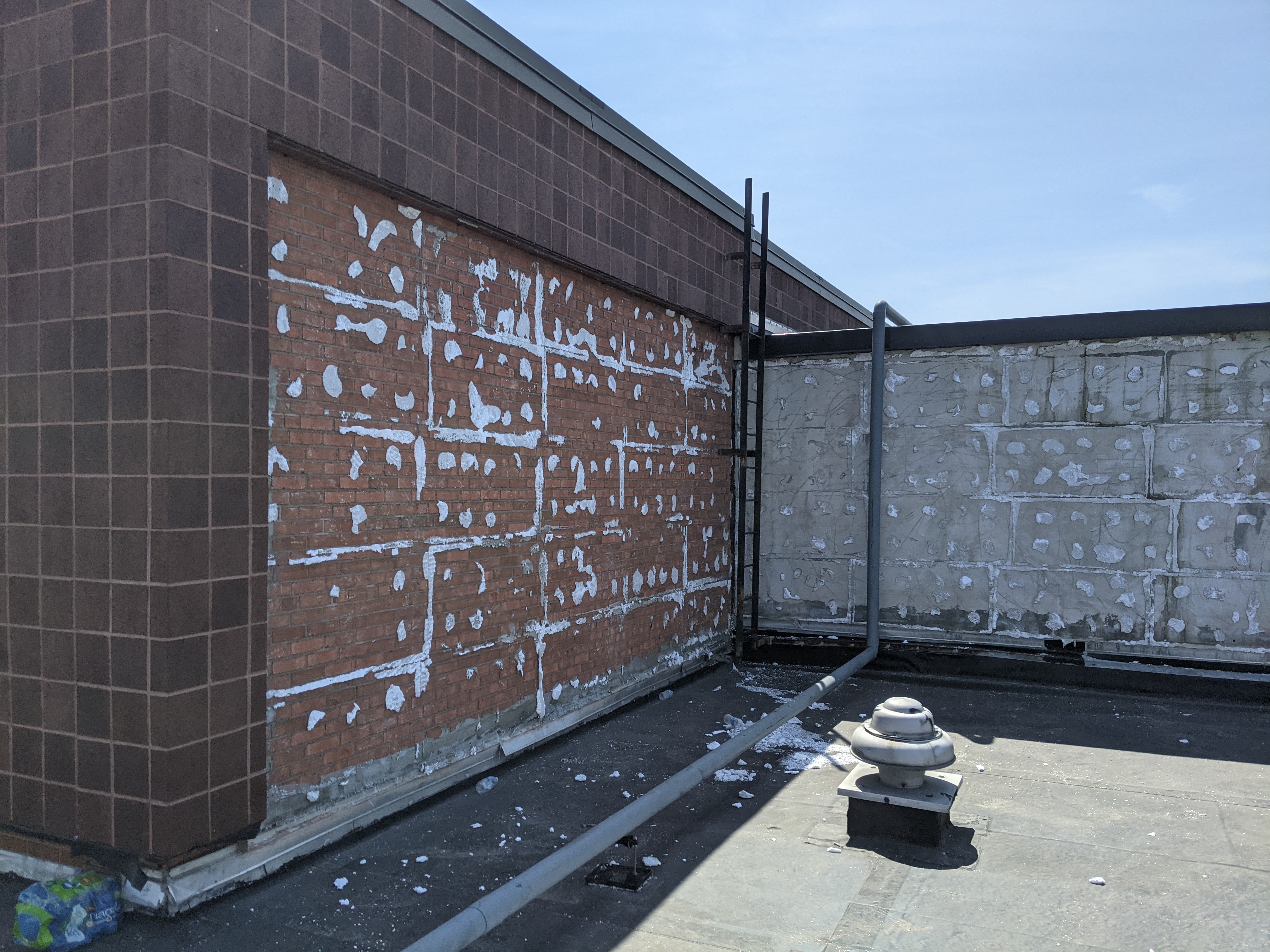 A rooftop with a partially exposed brick wall in the foreground and a concrete block wall in the background under a clear blue sky.