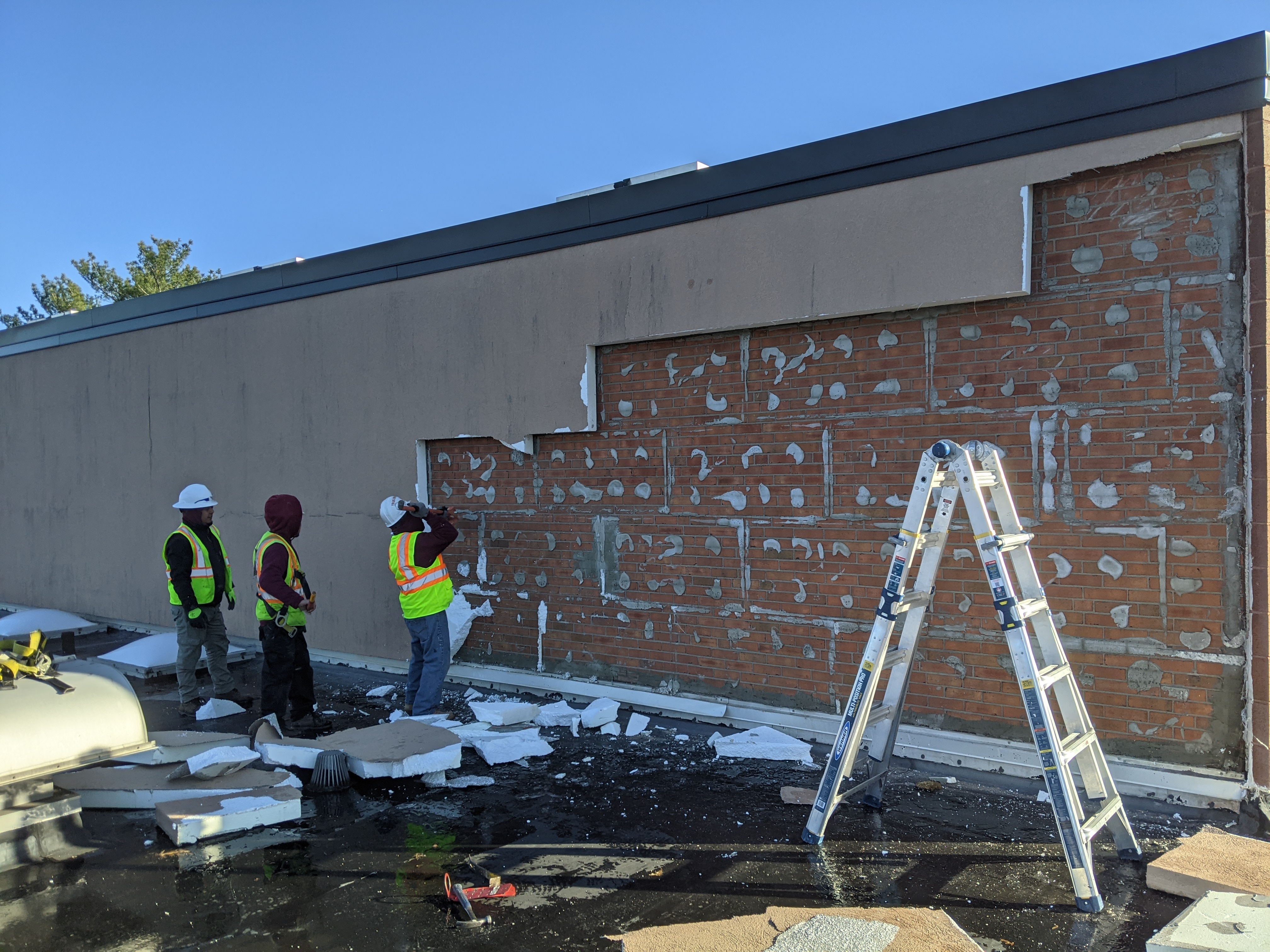 Workers in hard hats are removing siding from a brick building, with a ladder and debris in the foreground and a clear sky in the background.