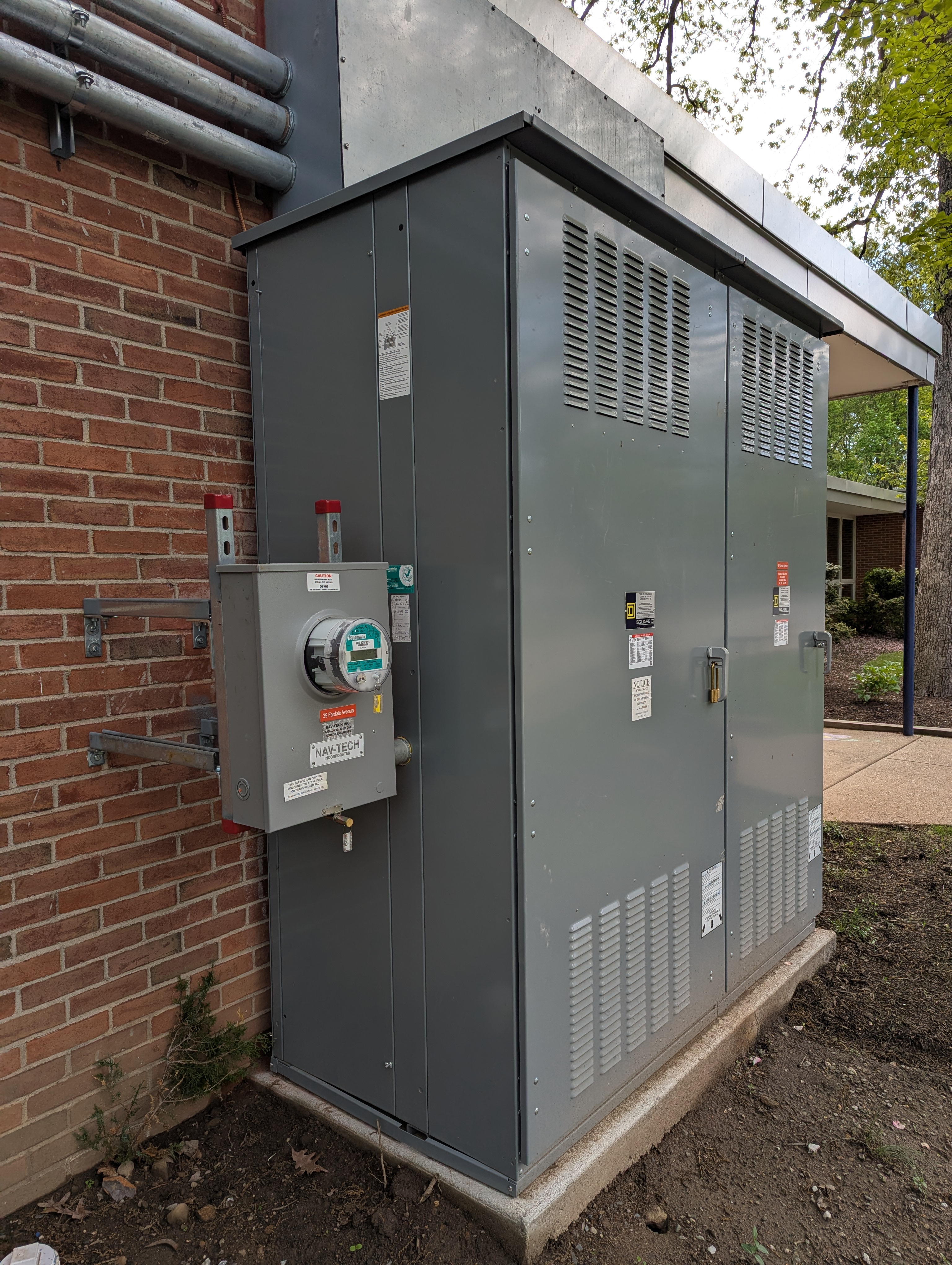 A large gray electrical transformer unit stands on a concrete pad in front of a brick building, with trees and a building visible in the background.