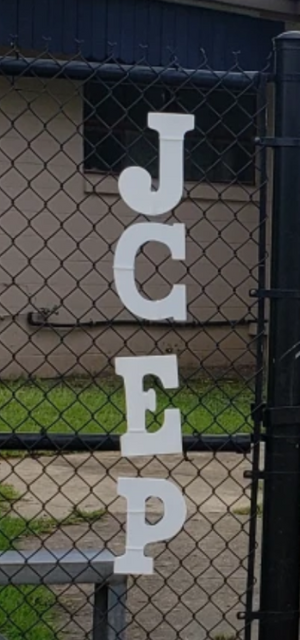 White letters spelling "JCEP" are attached to a chain-link fence in front of a building and grassy area.