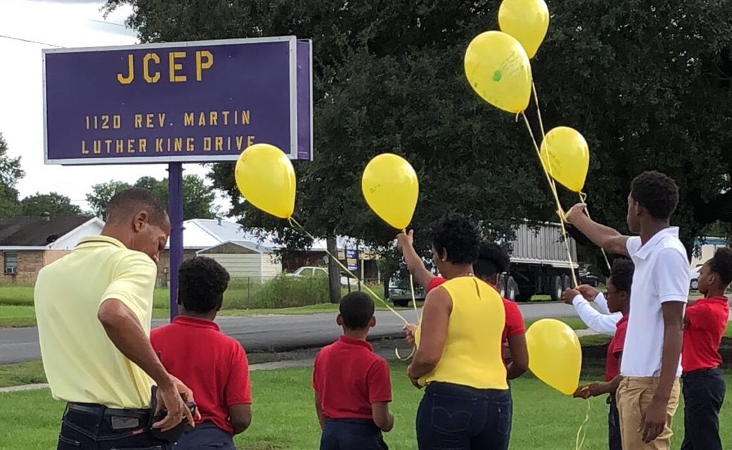 A group of children in red shirts and adults in yellow shirts stand in front of a sign that reads "JCEP 1120 REV. MARTIN LUTHER KING DRIVE" while holding yellow balloons.