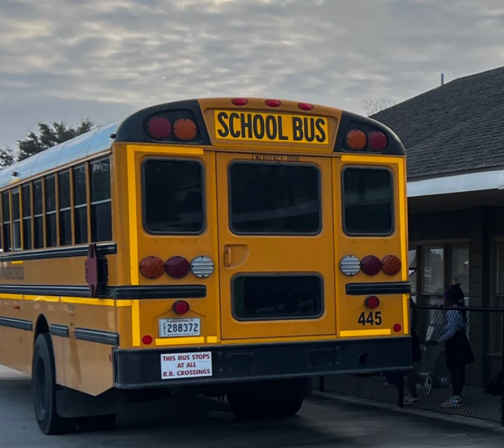 A yellow school bus with reflective tape is parked in front of a building with a dark roof, while a person stands near a fence in the background.