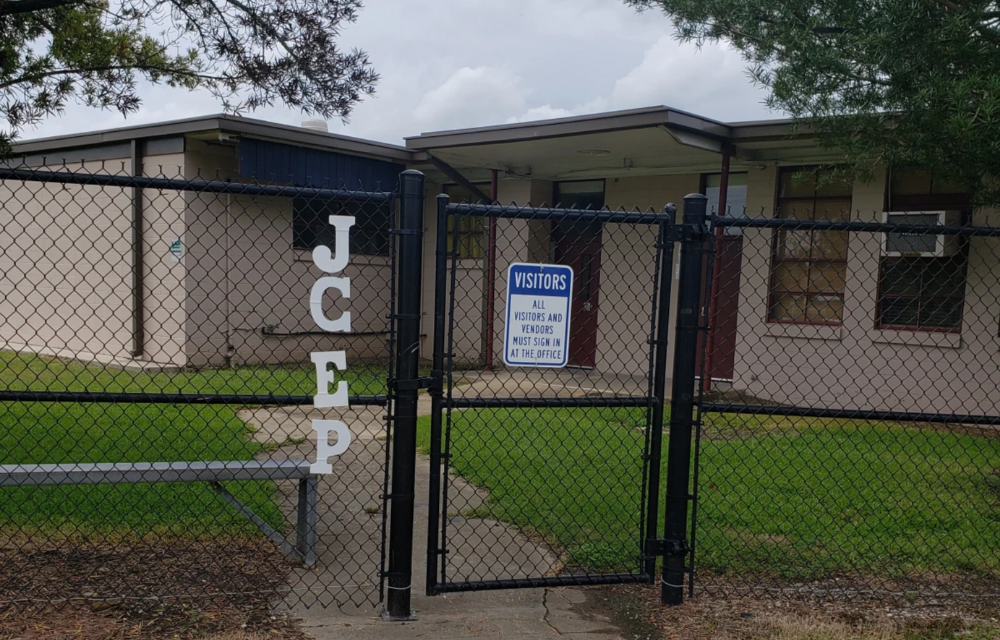A chain-link fence with a "JCEP" sign and a "Visitors" sign on a gate stands before a beige, single-story building with a dark roof and trees in the background.