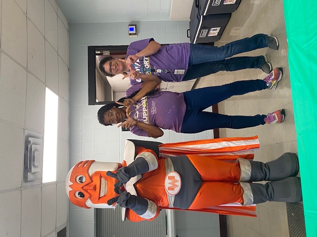 A mascot and two women pose in front of a hallway with storage bins.