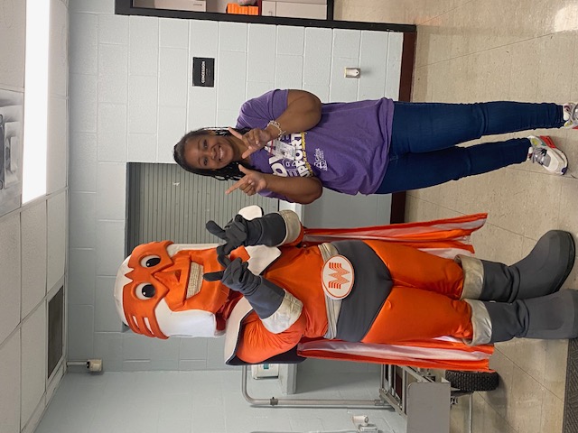 A person in a mascot costume stands next to a smiling woman in a purple shirt, with a hallway and lockers visible in the background.
