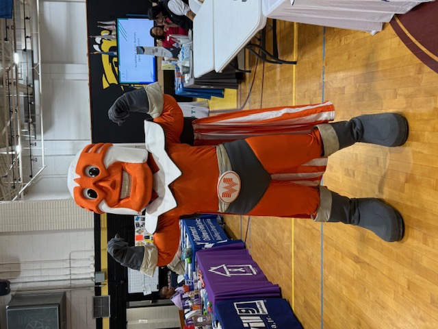 A mascot in an orange superhero costume flexes its muscles in the foreground, while a gymnasium with tables and people is visible in the background.