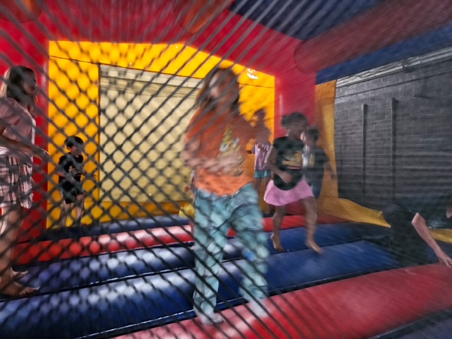 Children are joyfully jumping inside a colorful inflatable bounce house, with a blurry net in the foreground and a bright yellow and blue interior in the background.