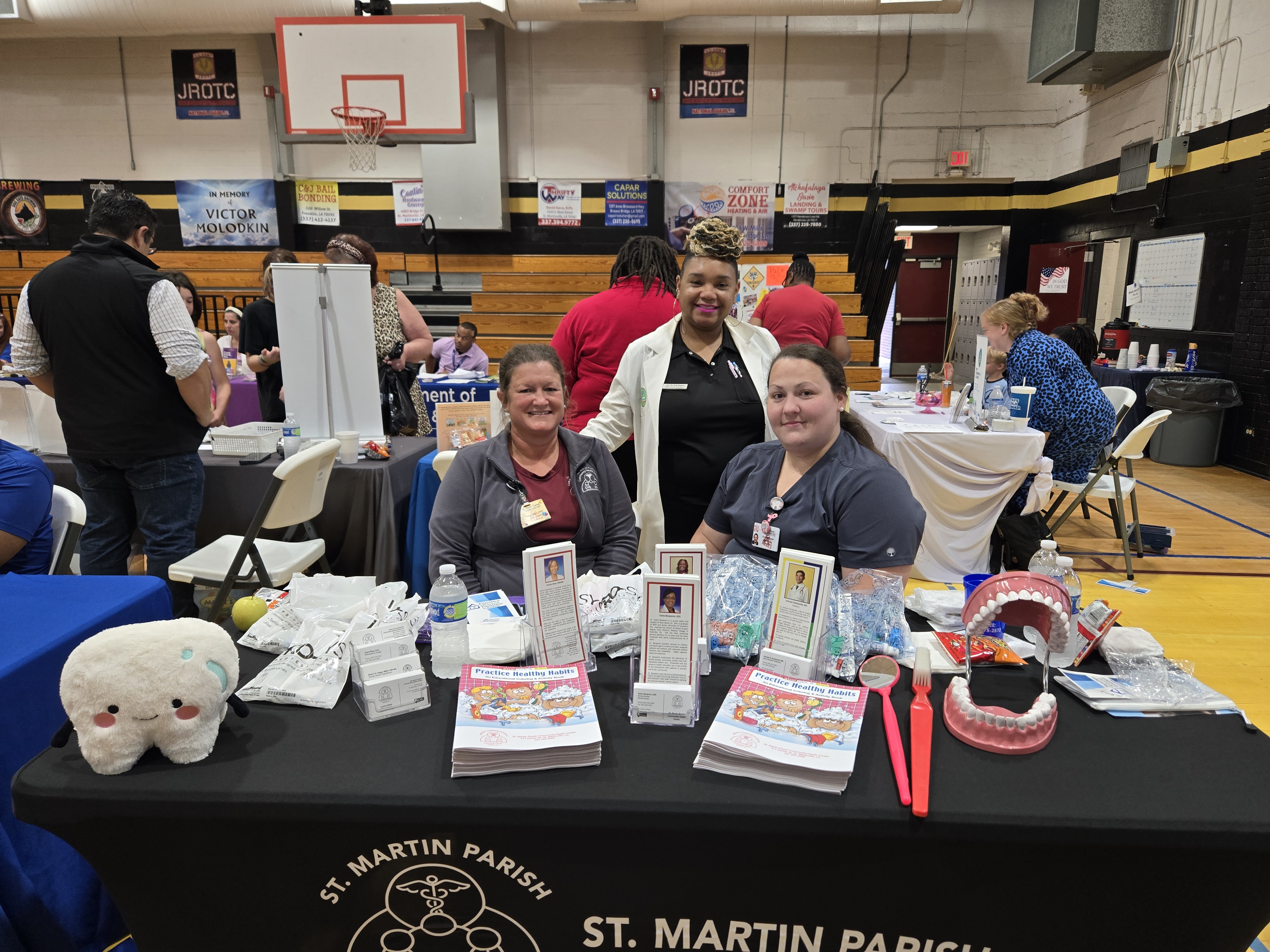 Three women stand behind a table with dental hygiene products in the foreground, while a gymnasium with various booths and people is visible in the background.