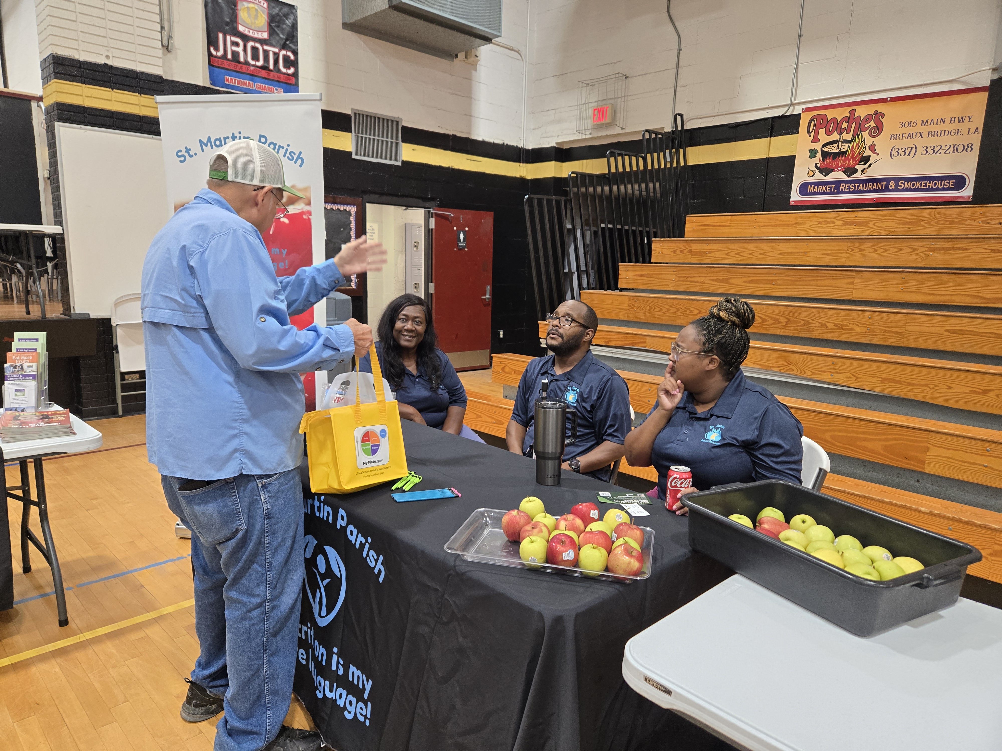In the foreground, a man in a blue shirt gestures towards a table with apples and a yellow bag, while in the background, three people sit behind the table with bleachers and a JROTC banner visible.