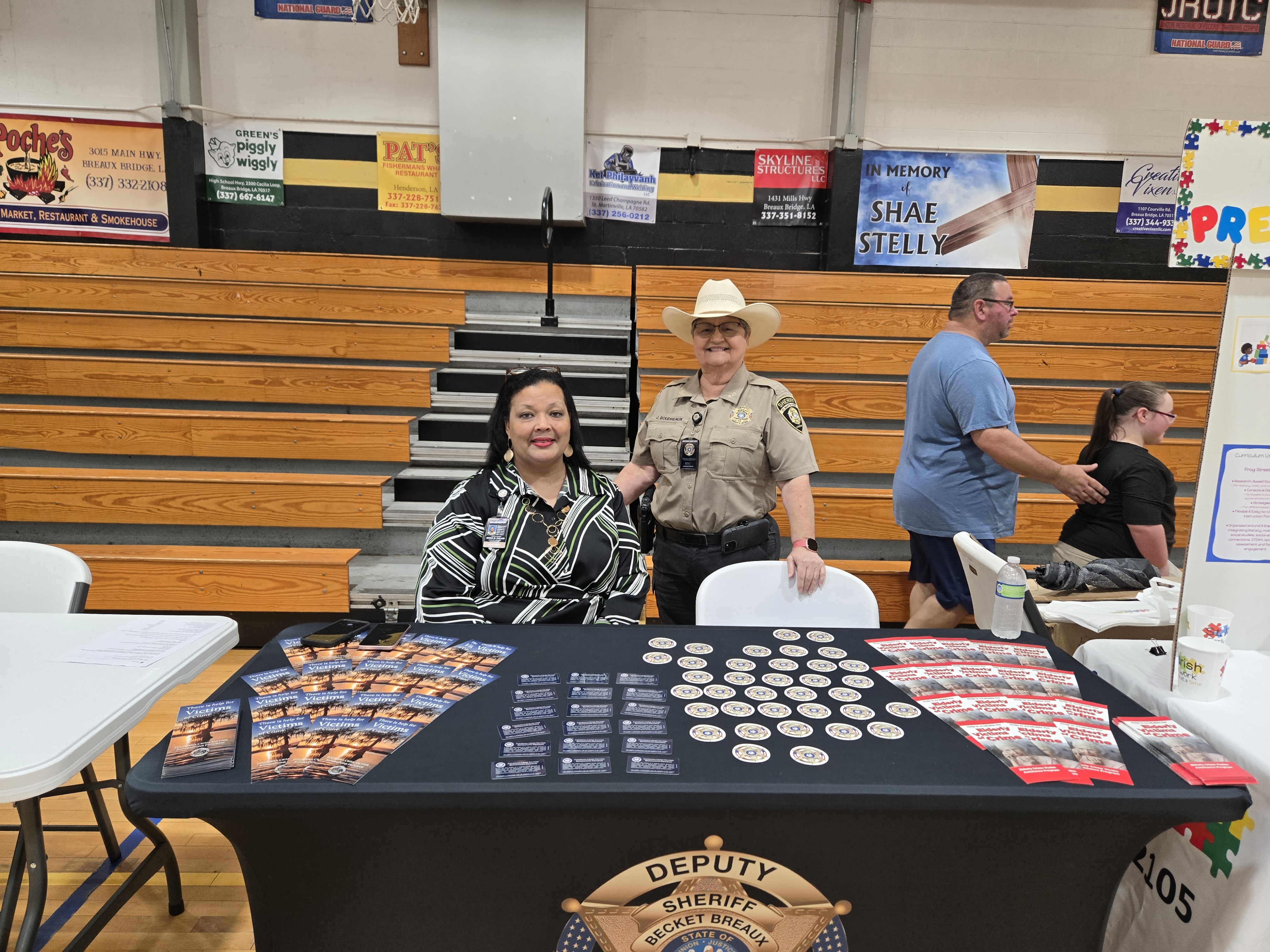 Two women, one in a sheriff's uniform, stand behind a table displaying promotional materials, with bleachers and various signs visible in the background.