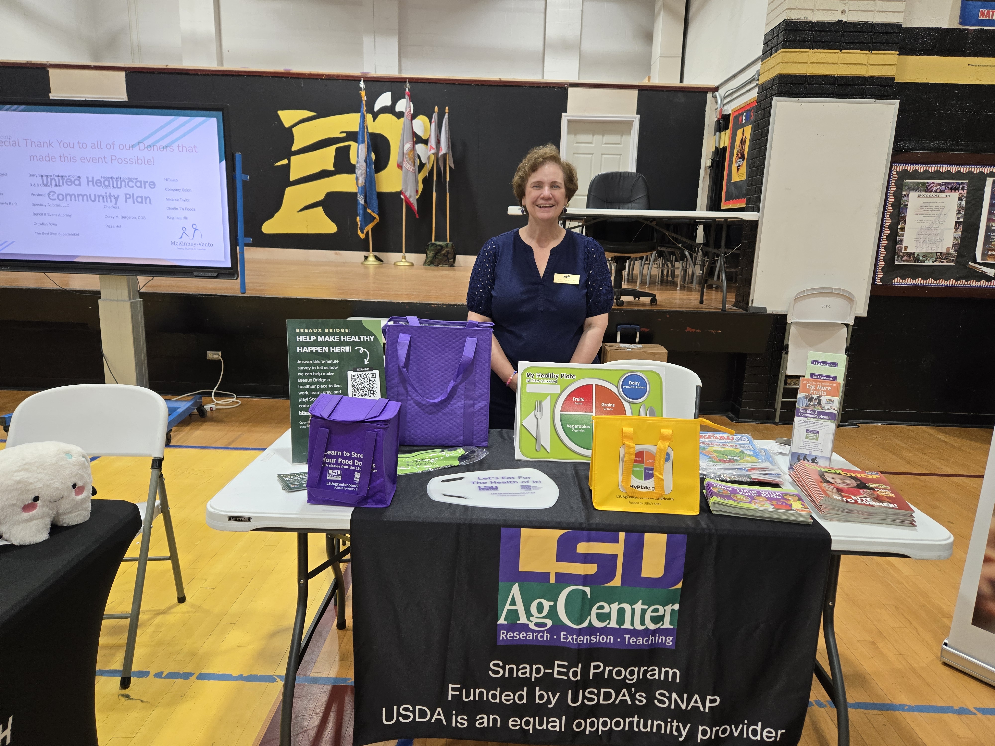 A woman stands behind a table displaying LSU AgCenter materials, with a gymnasium in the background.