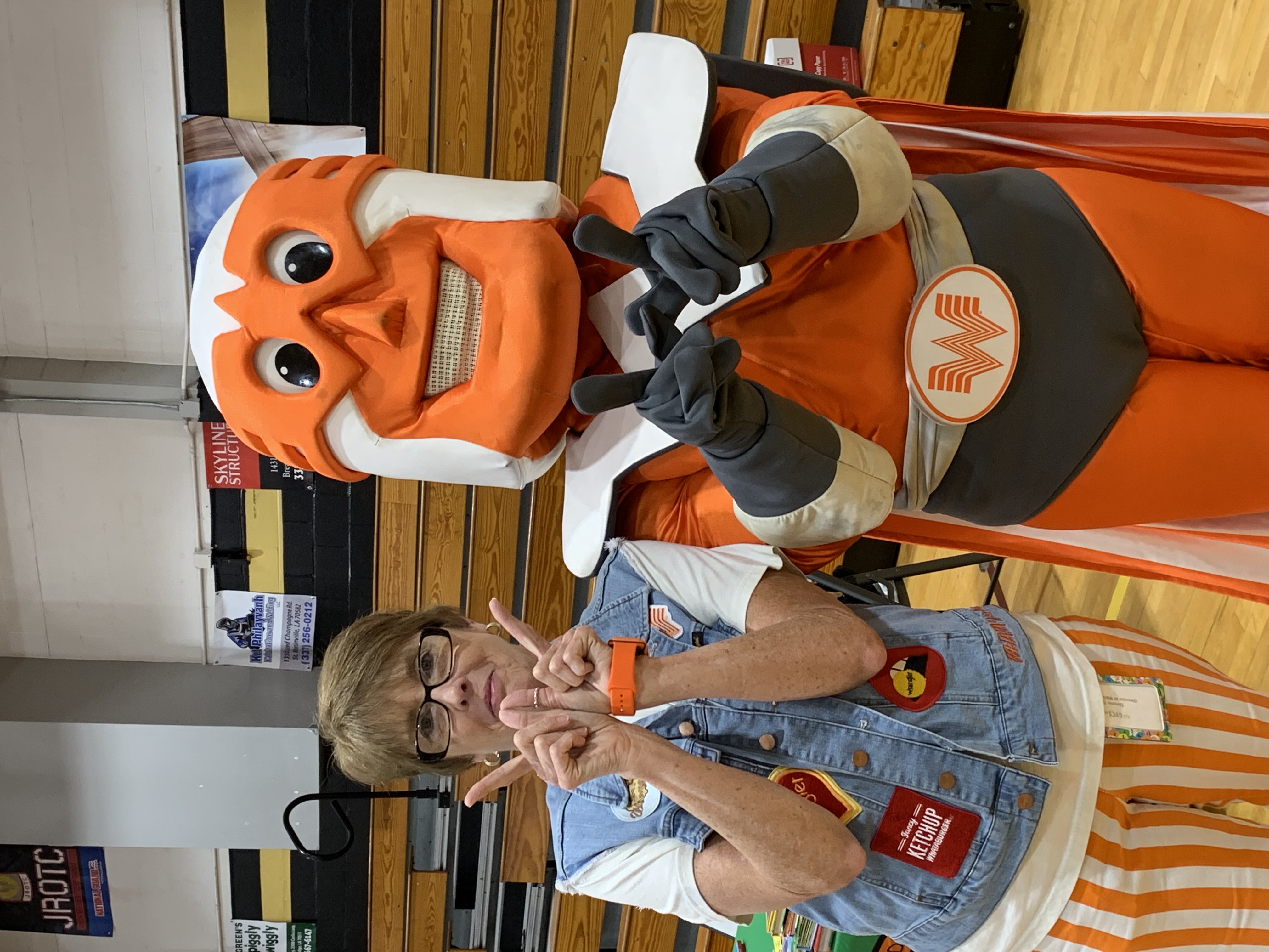 A woman in a denim vest and striped pants poses with a mascot in an orange superhero costume, with a gymnasium in the background.
