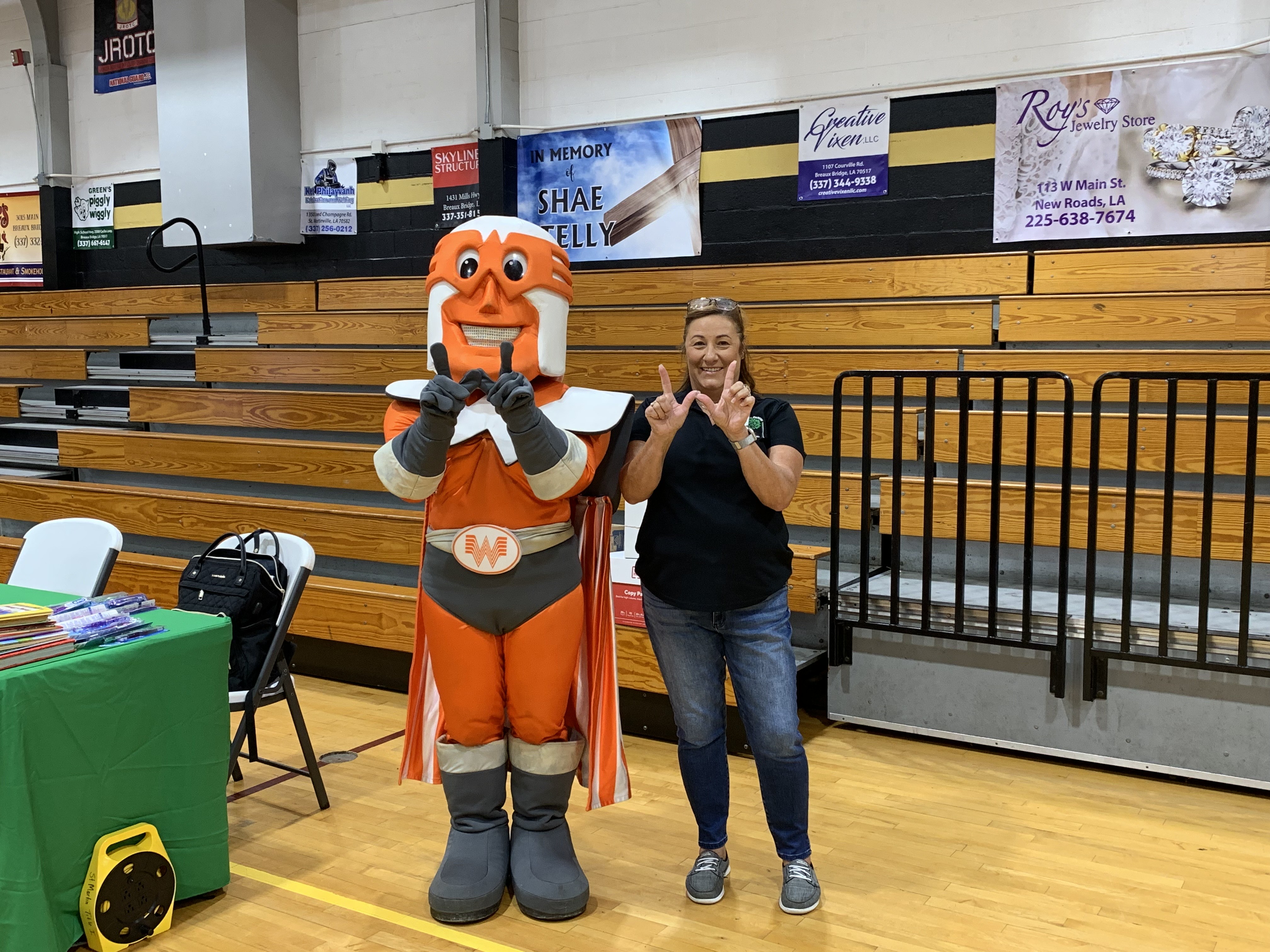 A mascot and a woman pose in front of bleachers and banners in a gymnasium.