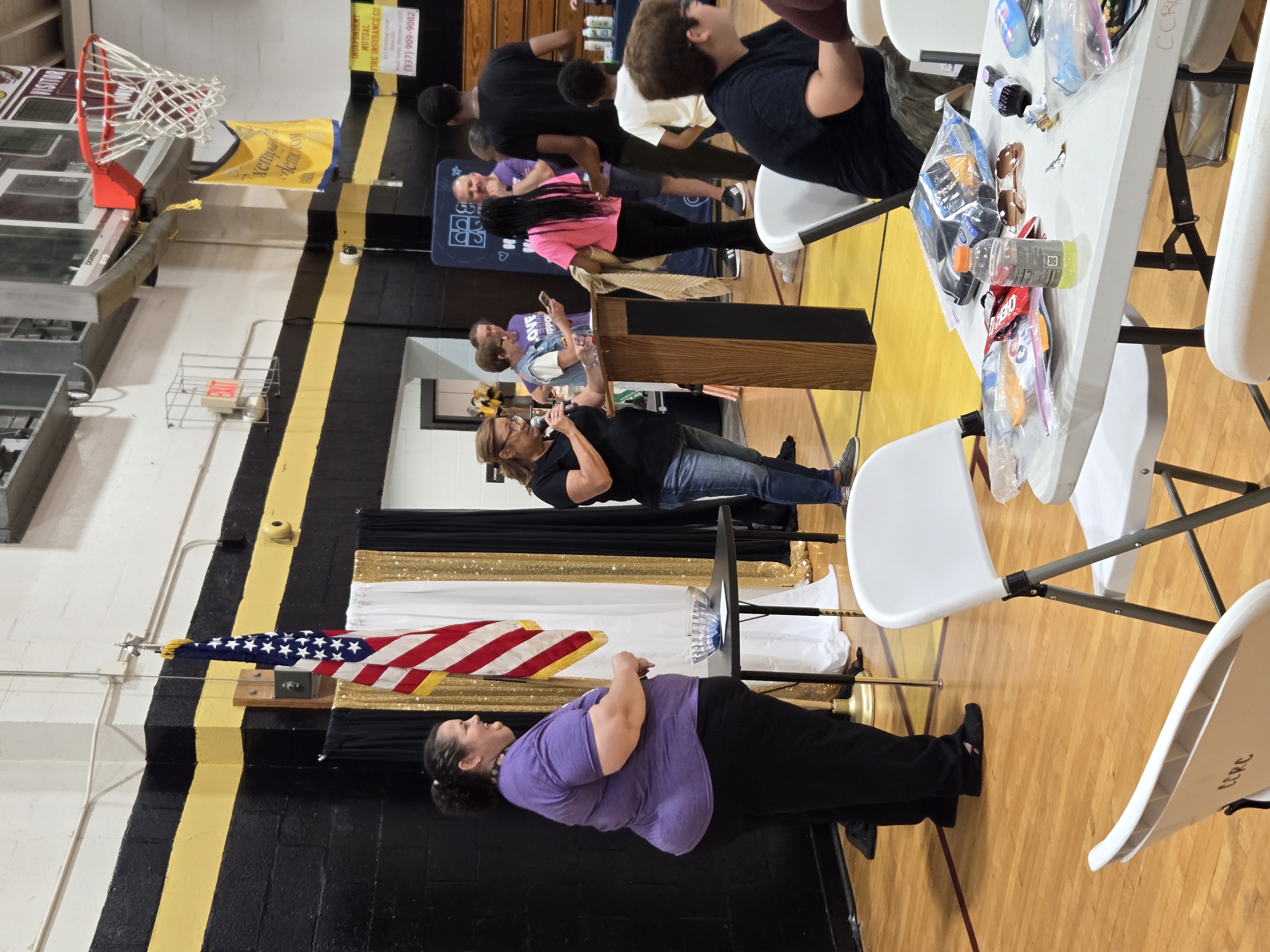 In the foreground, white folding tables are set up with various items, while in the background, people gather in a gymnasium with a basketball hoop and an American flag.