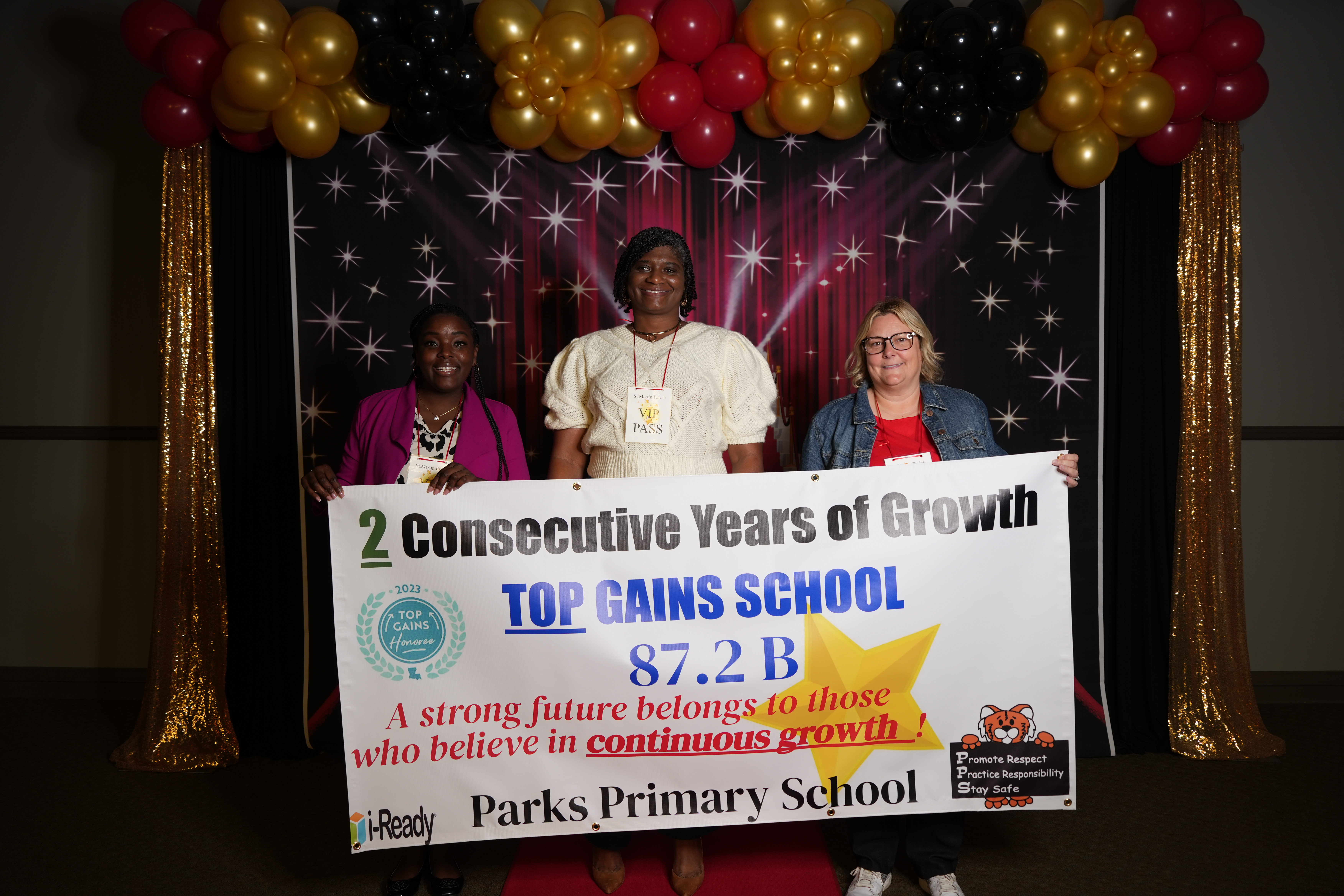 Three women stand behind a banner celebrating "2 Consecutive Years of Growth" for "TOP GAINS SCHOOL" at "Parks Primary School," with a sparkly backdrop and balloons.