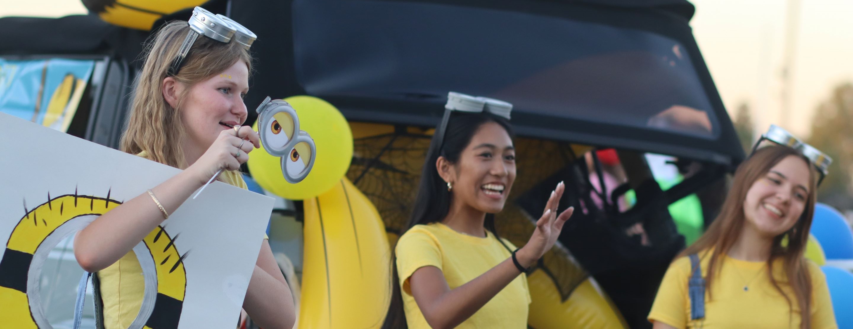 Three young women in yellow shirts and goggles stand in front of a car trunk decorated with Minion-themed balloons and a life preserver.