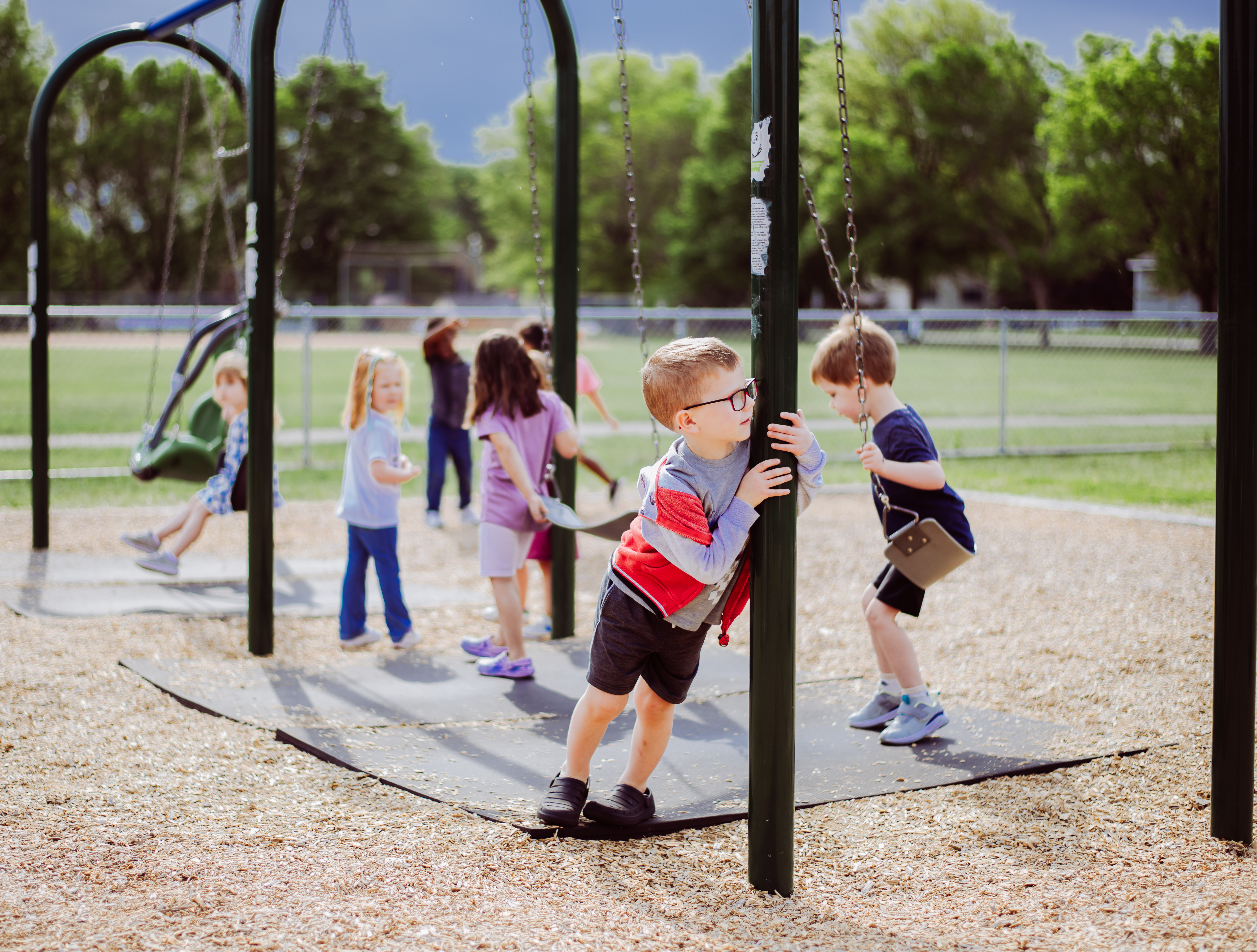 Children play on swings and around a swing set in a park with trees and a fence in the background.