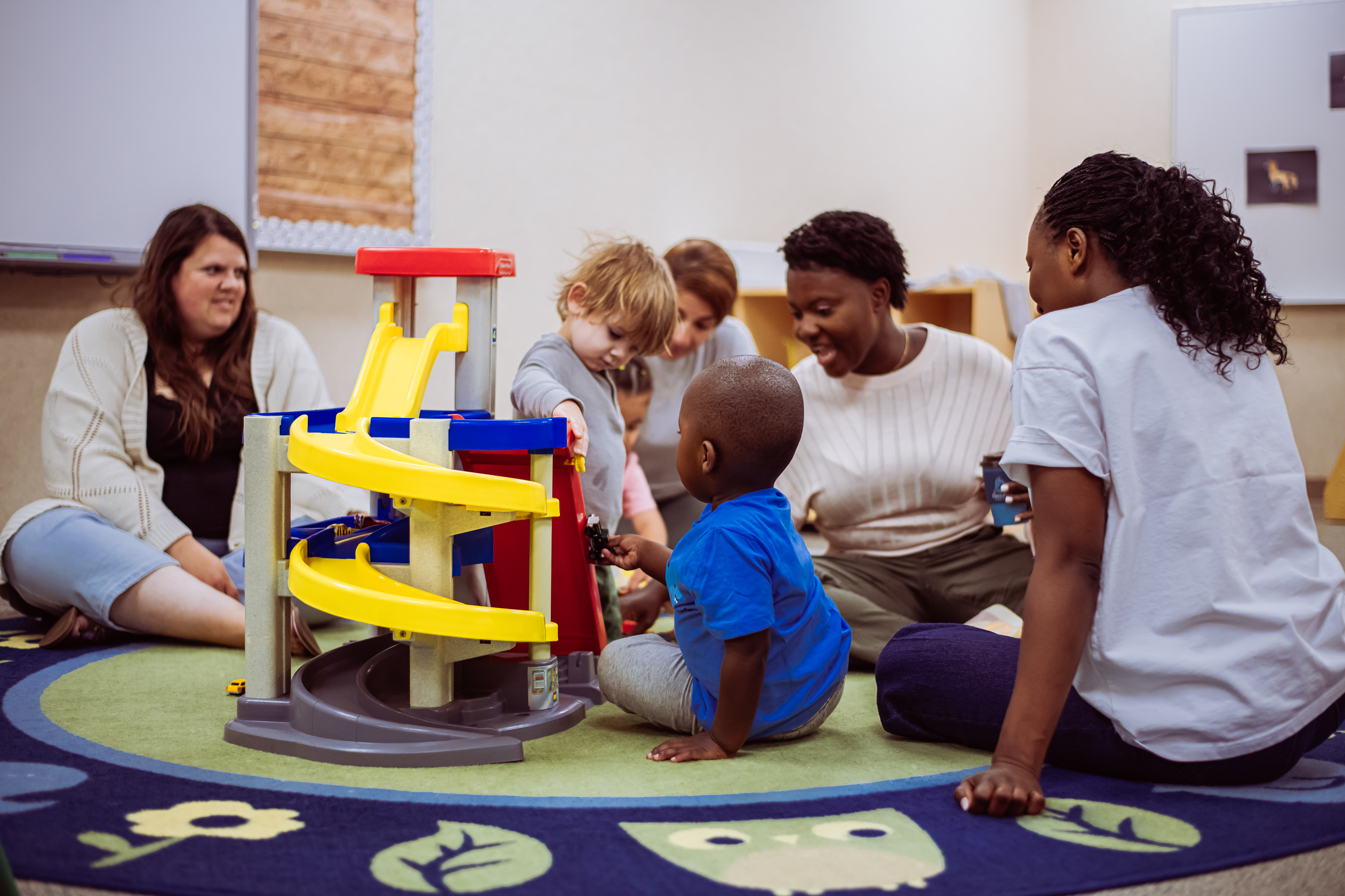 Children and adults are gathered around a colorful toy car ramp on a patterned rug, with a plain wall and shelves in the background.
