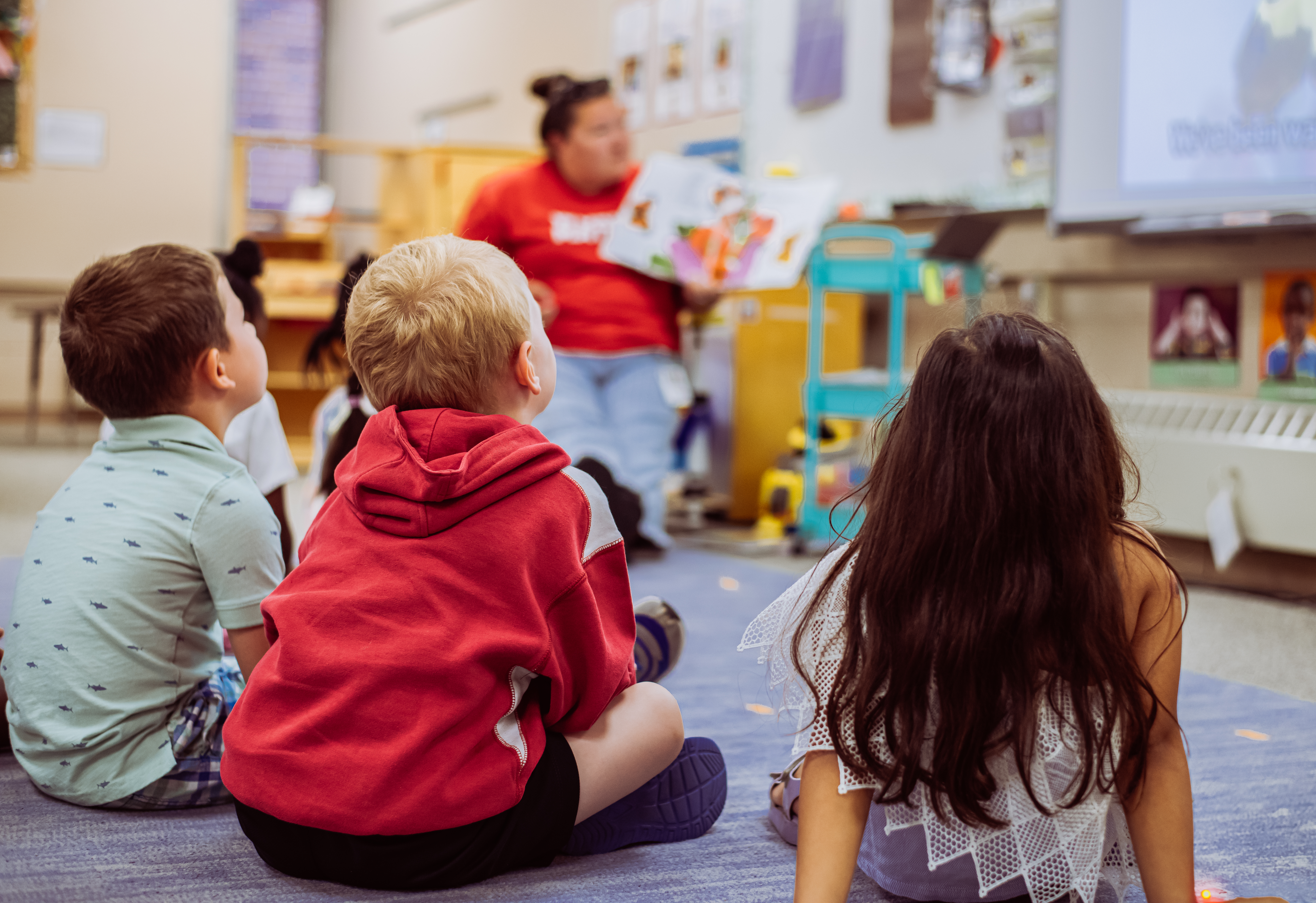 Young children sit on a blue rug in the foreground, attentively watching a teacher read a book in the background.