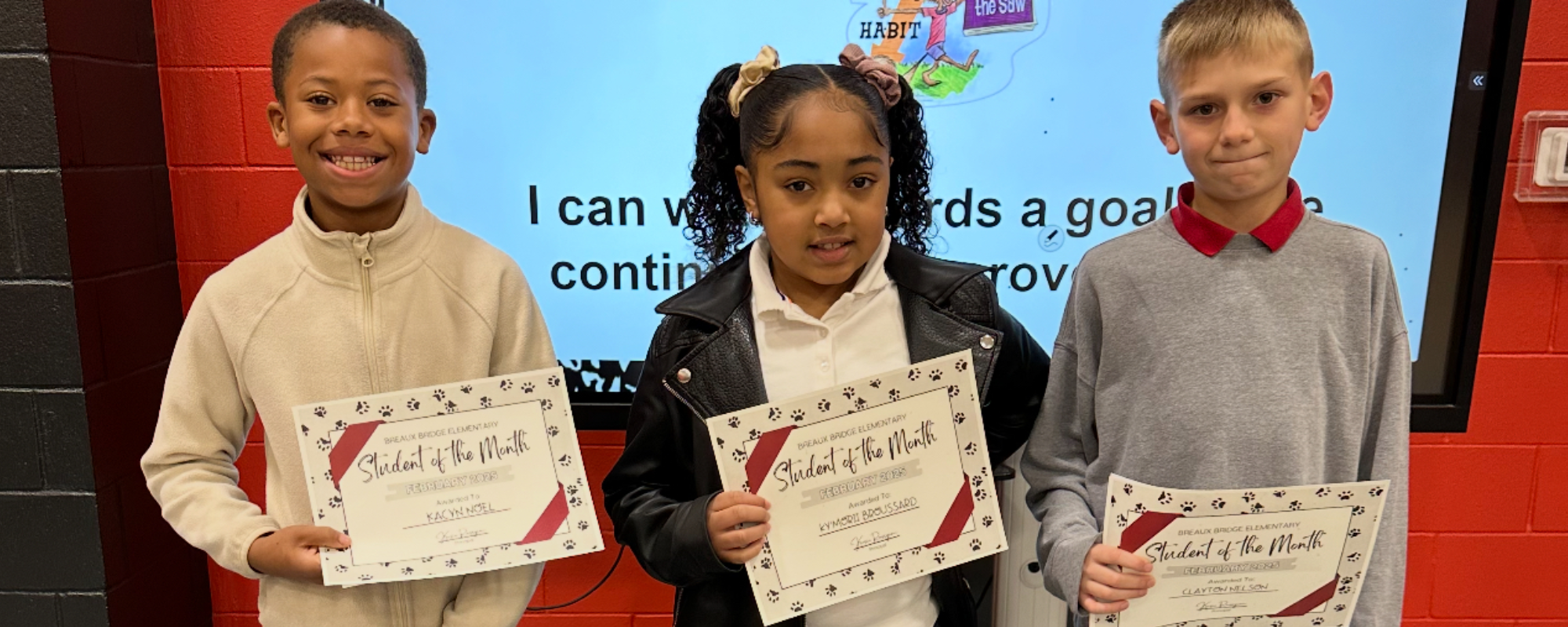 Three smiling children hold "Student of the Month" certificates in front of a classroom screen displaying text and colorful graphics.