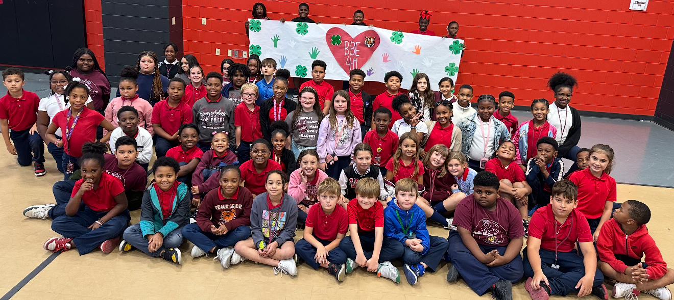 A large group of children in red shirts are sitting on the floor in the foreground, while a banner with shamrocks and hearts hangs on the red wall in the background.