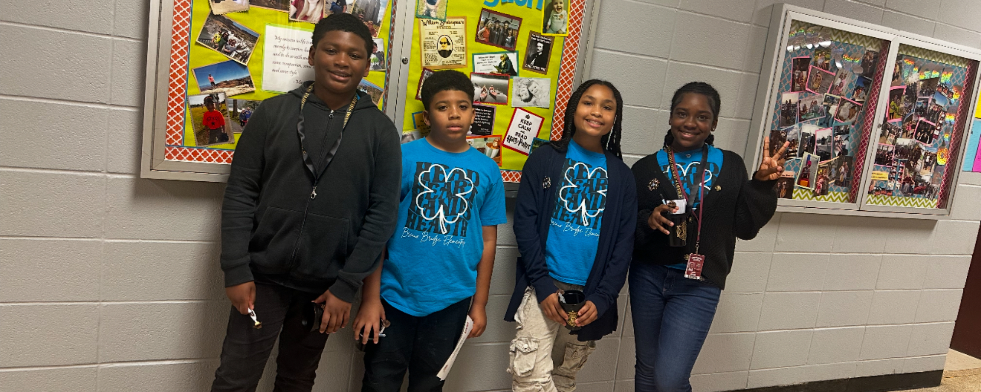 Four young people stand in front of a bulletin board and a display case in a hallway.