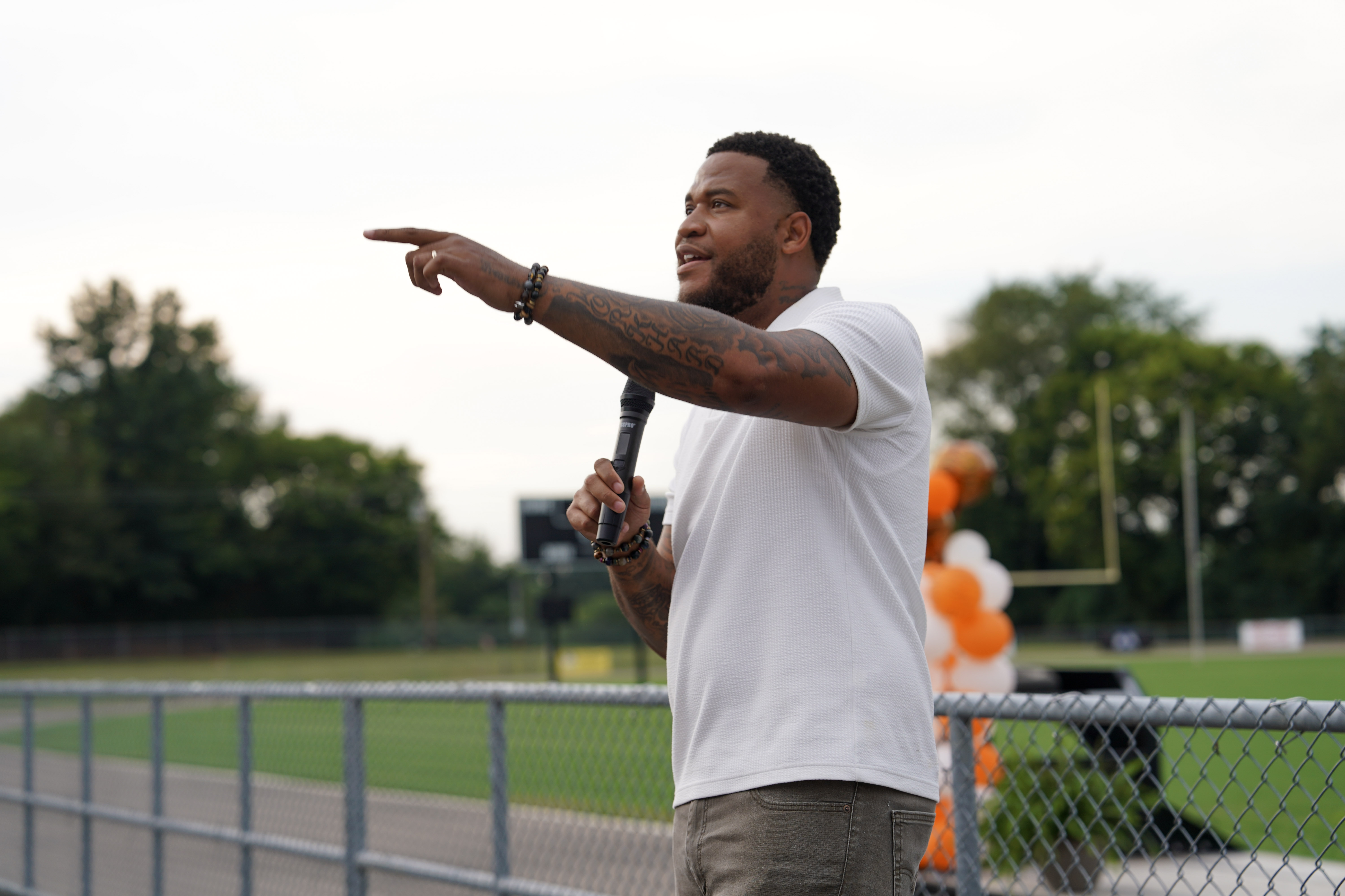 A man holding a microphone gestures and points while speaking outdoors near a sports field, with trees and orange and white balloons visible in the background.