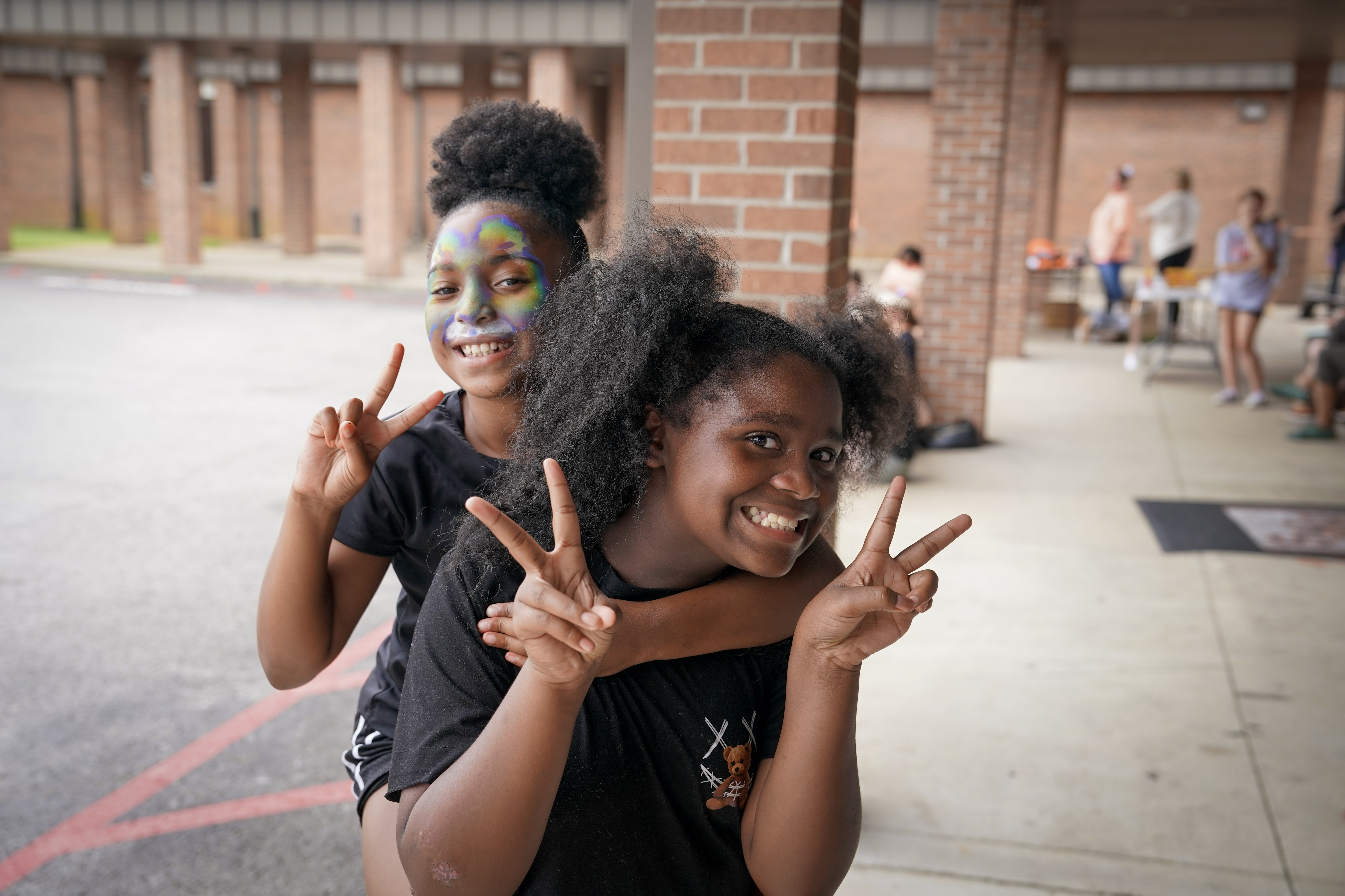 Two smiling children pose outside a building. One, with colorful face paint, gives a piggyback ride and both hold up peace signs with their fingers. Other people are visible in the background under a covered walkway.