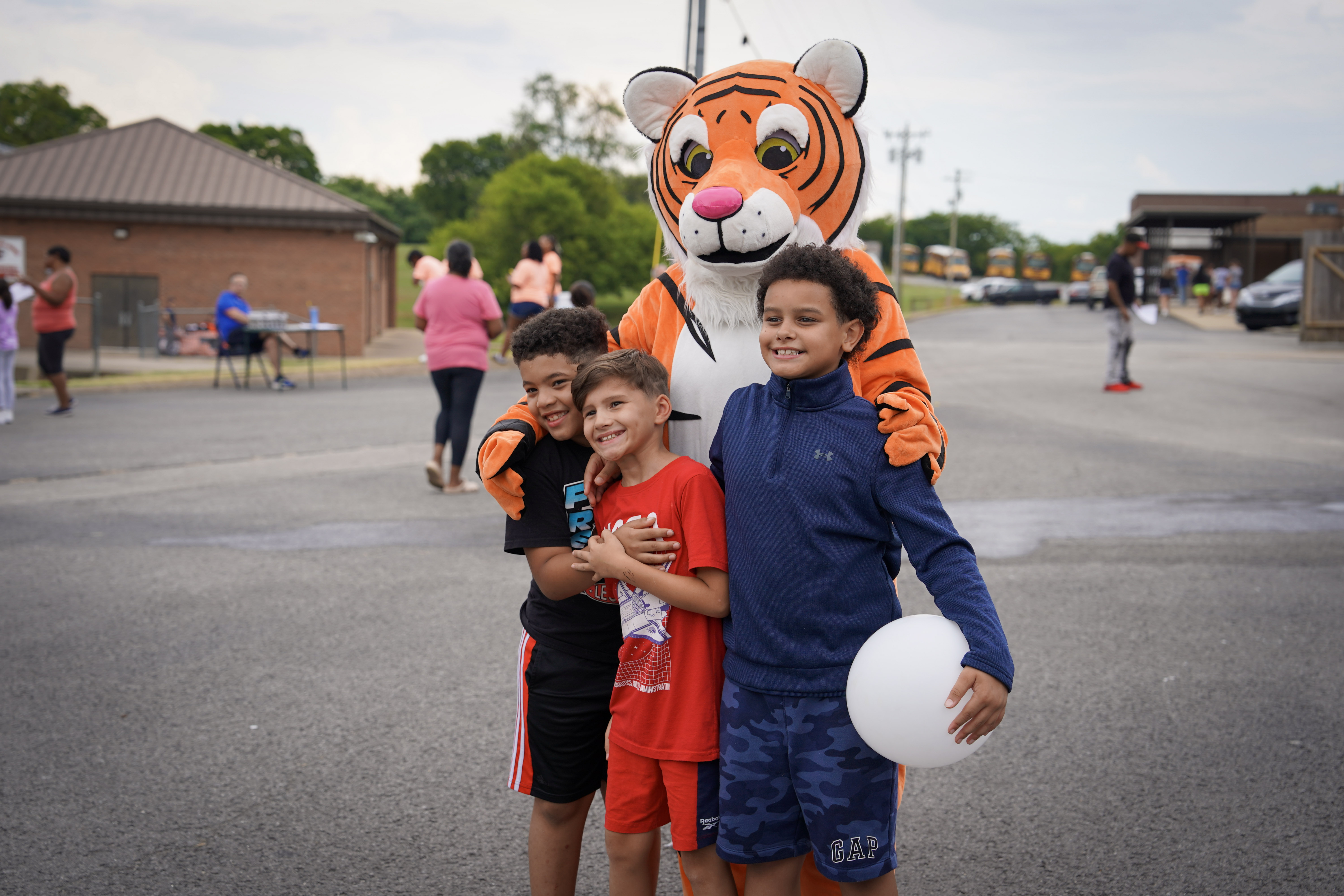 Three smiling boys pose together outdoors, with a person in a friendly tiger mascot costume standing behind them, arms around their shoulders. One boy holds a white balloon. Other people and buildings are visible in the background.