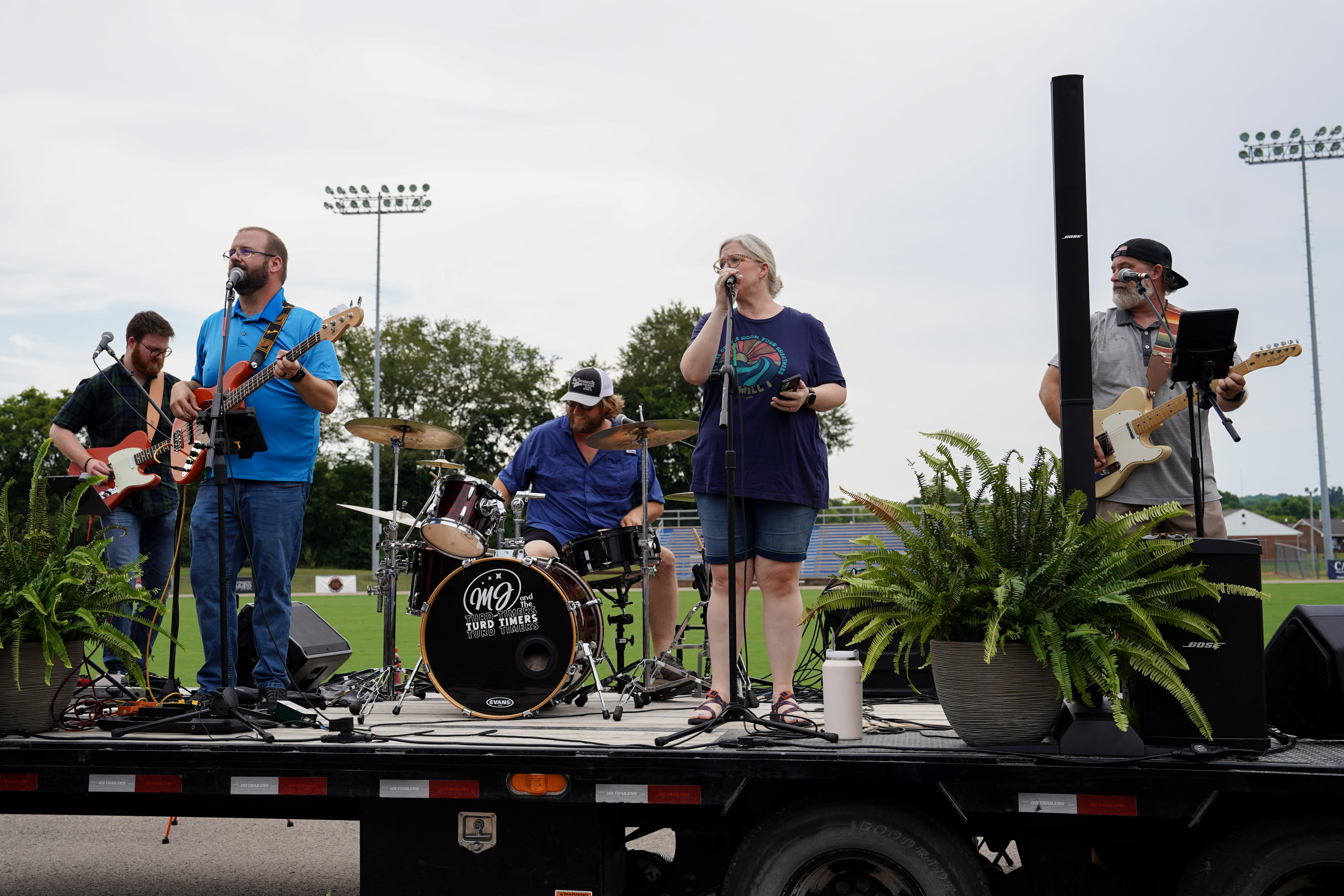 A five-member band performs on an outdoor stage. There are two guitarists, a drummer, a singer with a microphone, and another musician with a microphone and tablet. Potted plants decorate the stage. Stadium lights are visible in the background.